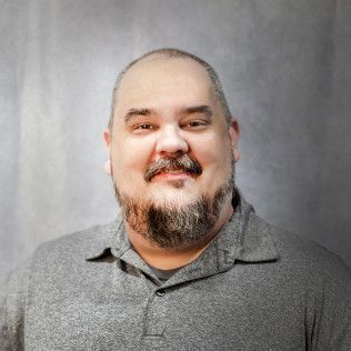 A man with a beard is wearing a grey shirt and smiling for the camera.