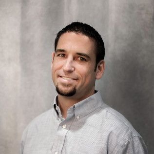 A man with a beard is wearing a grey shirt and smiling for the camera.