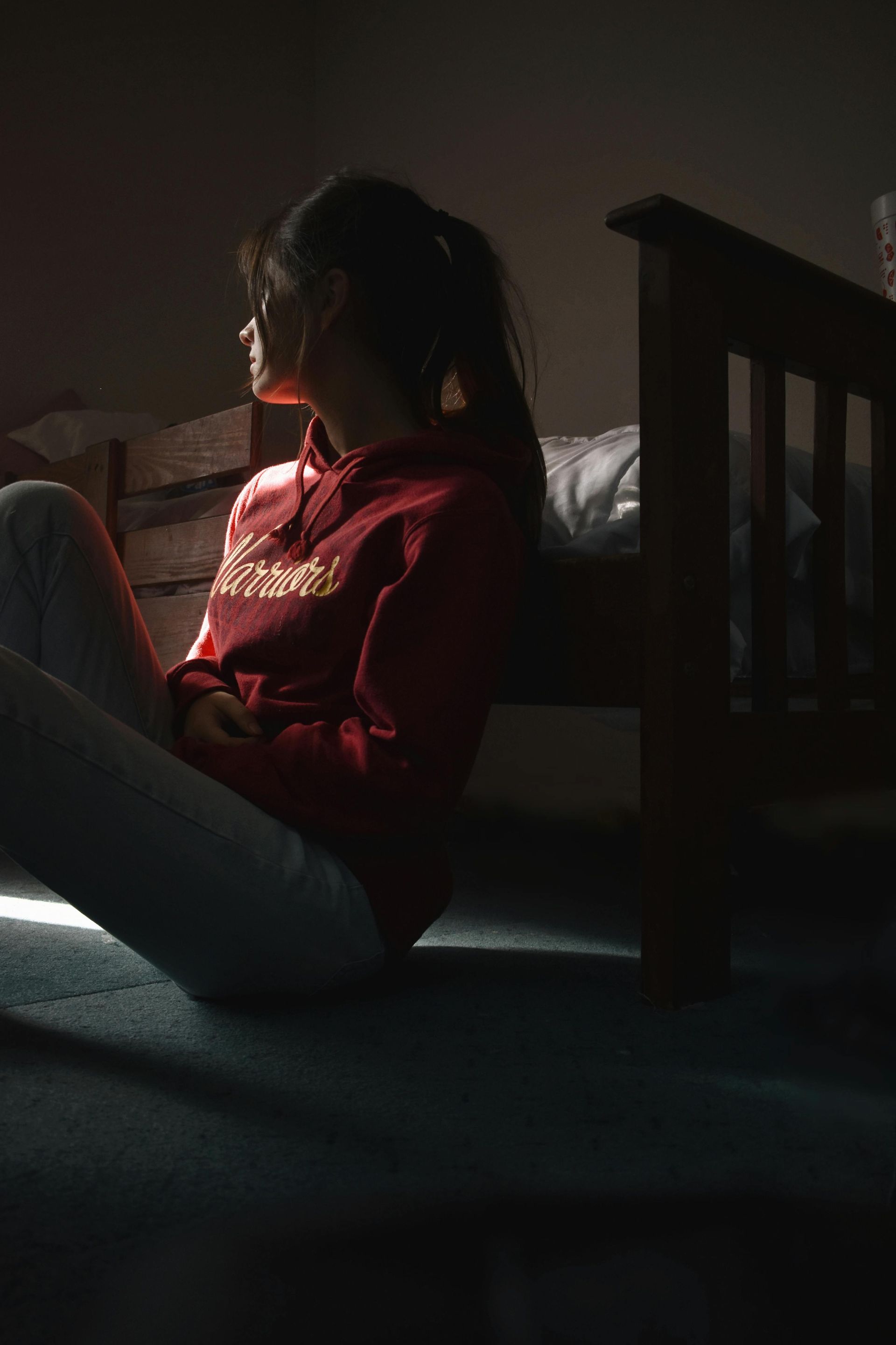 A woman in a red sweatshirt is sitting on the floor in front of a bed.