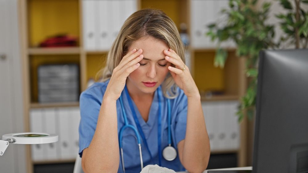 A nurse is sitting at a desk with her hands on her head.