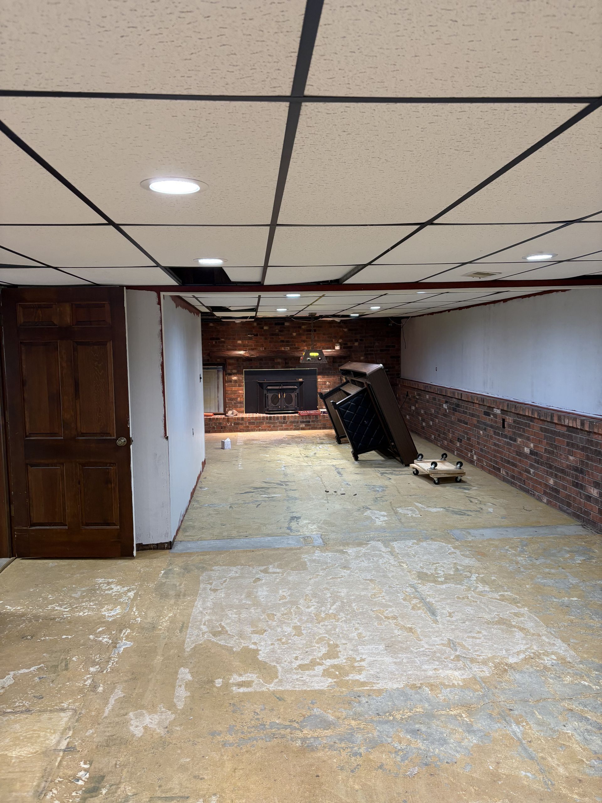 Basement renovation in progress. Exposed brick wall, brown door, ceiling tiles, debris on floor, furniture.