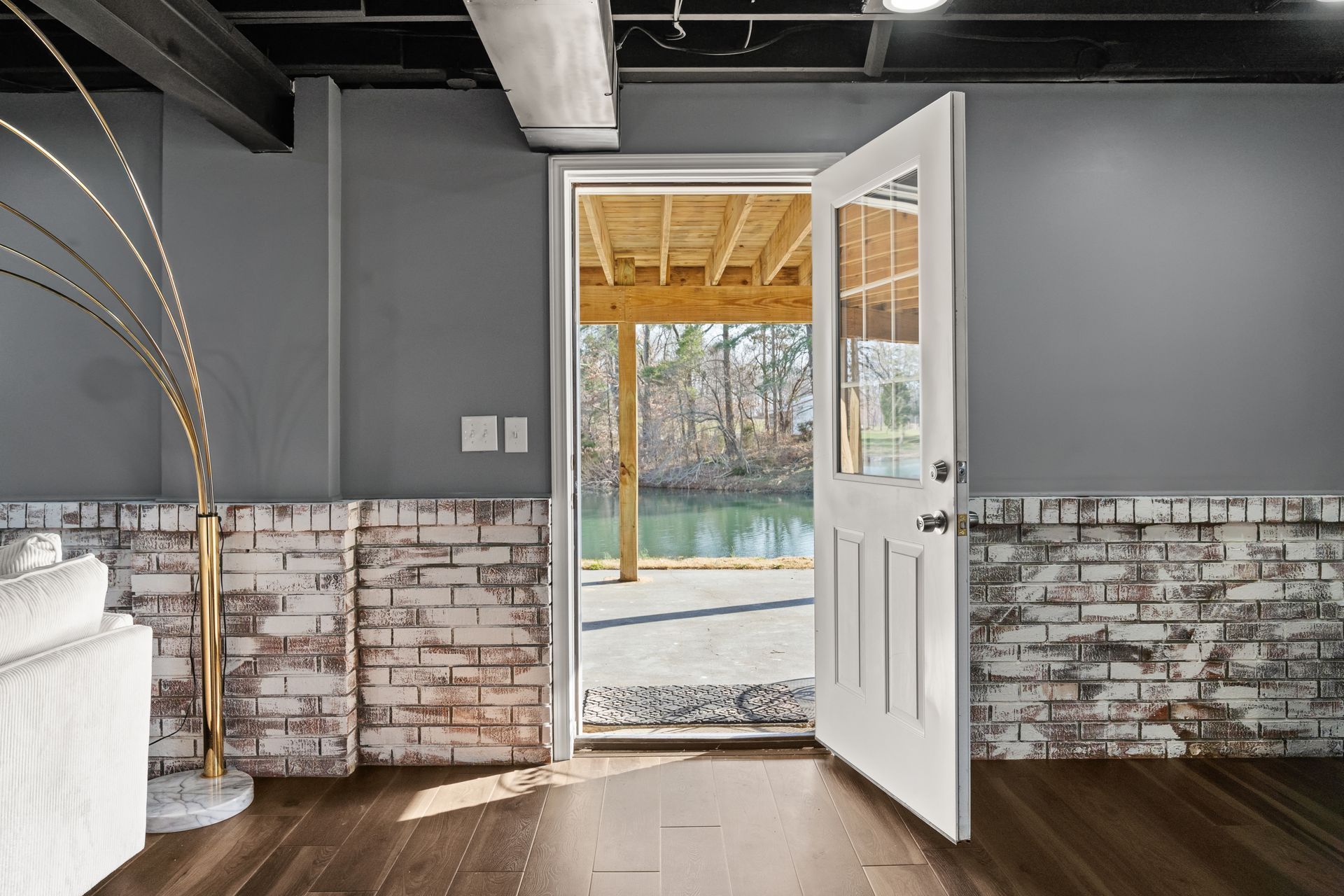 Basement interior with open white door leading to a deck overlooking water. Gray walls, exposed beams, brick accents.