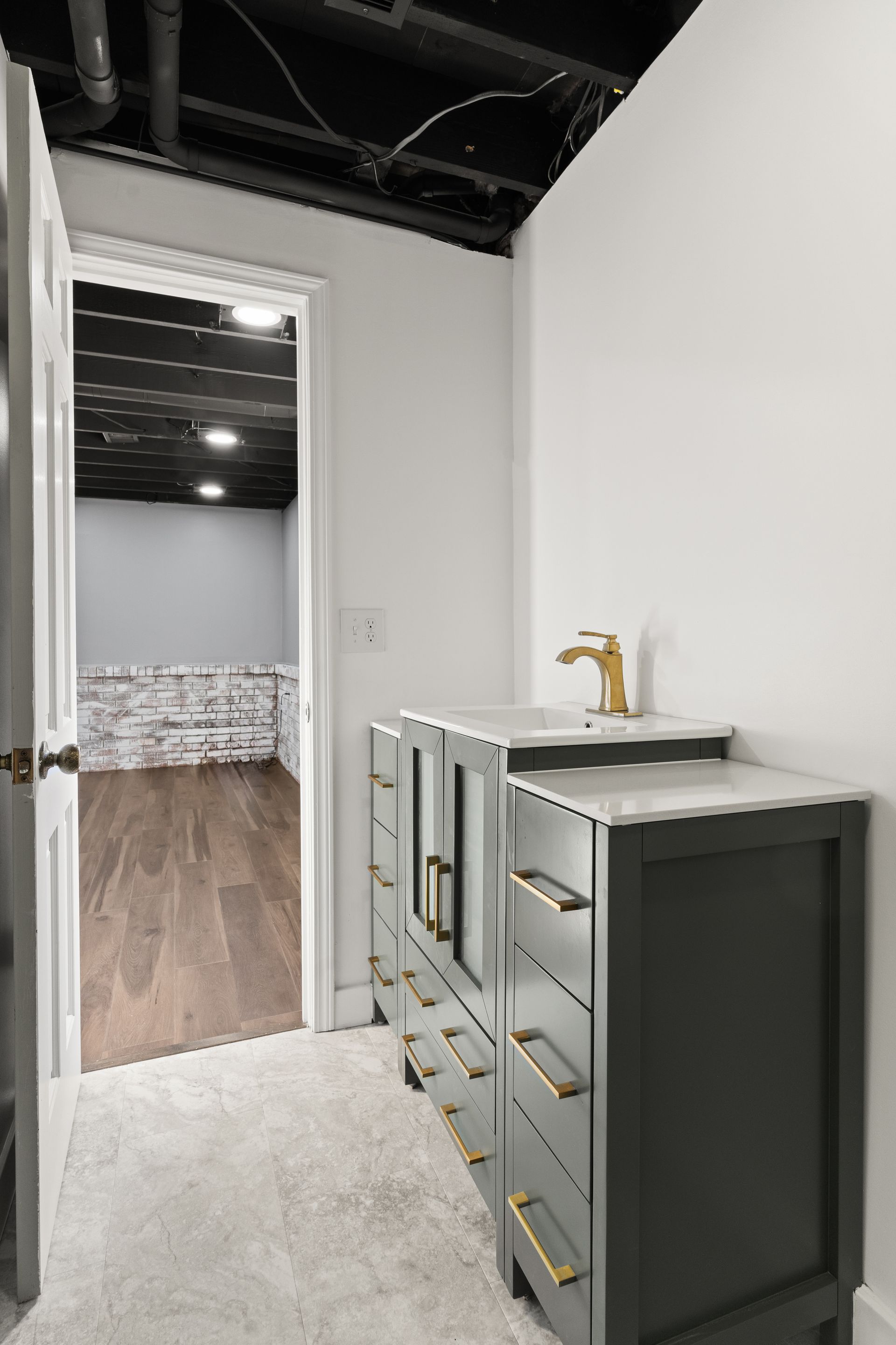 Bathroom with gray vanity, gold faucet, and doorway to a room with exposed brick and hardwood floors.