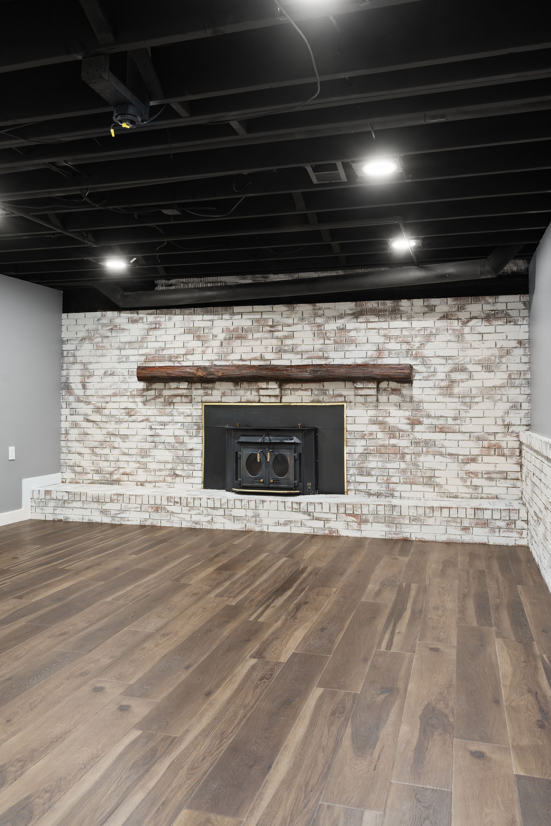Finished basement with fireplace and dark wood flooring, black ceiling. Brick accent wall and light fixtures.