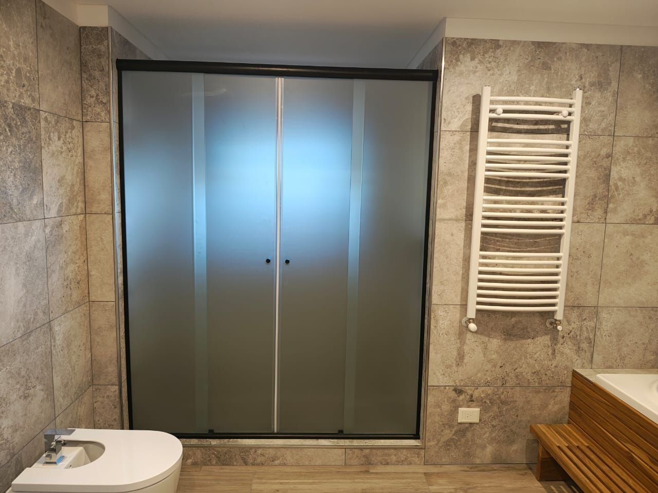 Baño moderno con pared de ducha de cristal, colores neutros y cama visible más allá.