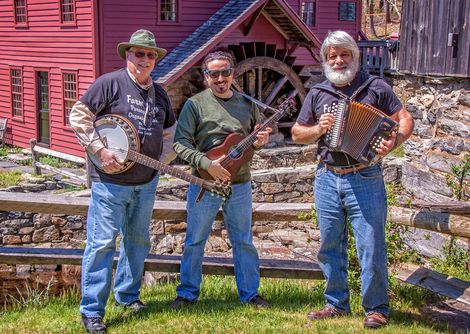 Doc Wood holding a banjo, Ian Reyes holding a bass guitar, Jon Dember holding an accordion in front of a mill.