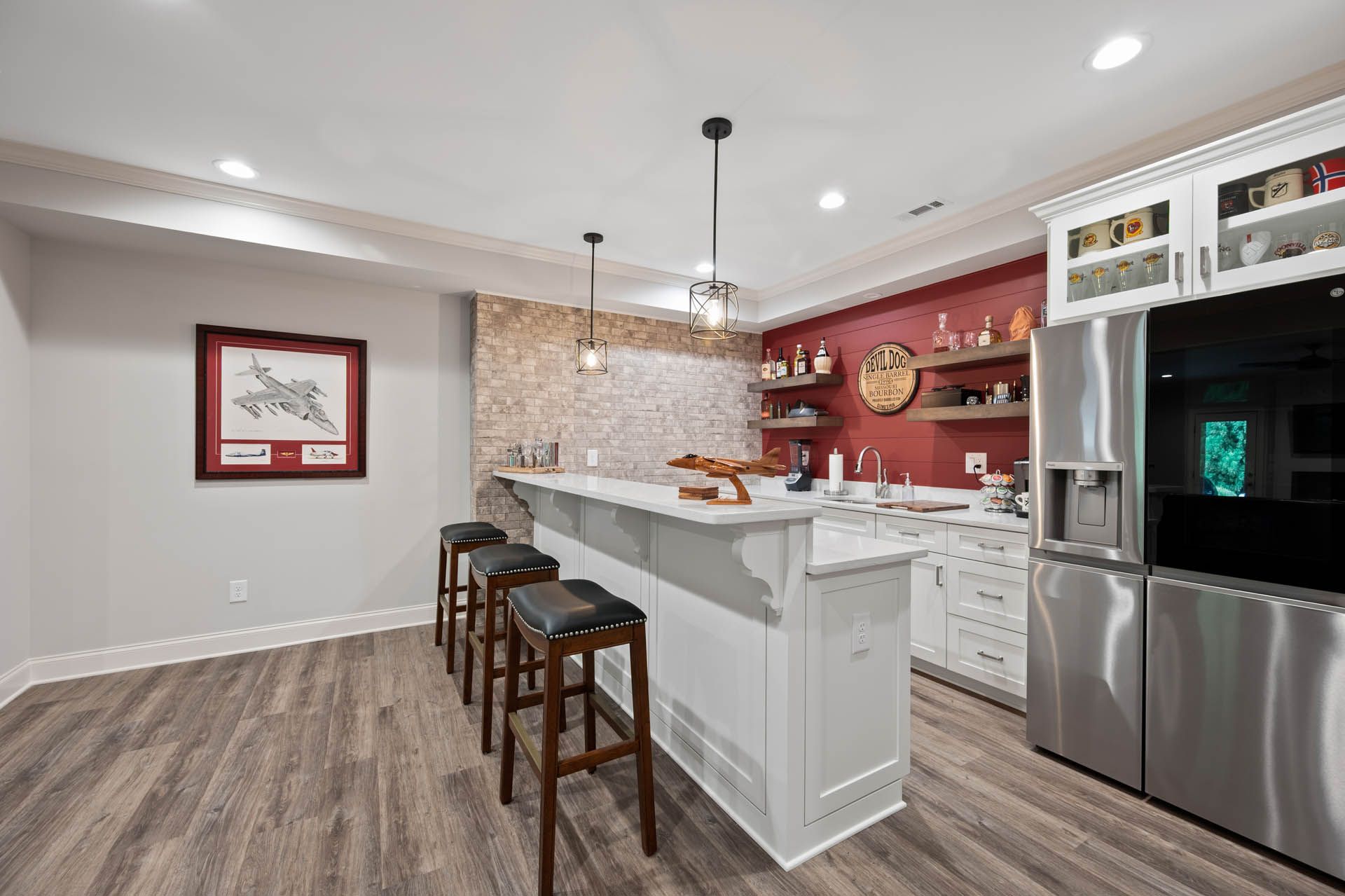 A well-lit basement bar with a white island, bar stools, and a stainless steel refrigerator against a red accent wall.