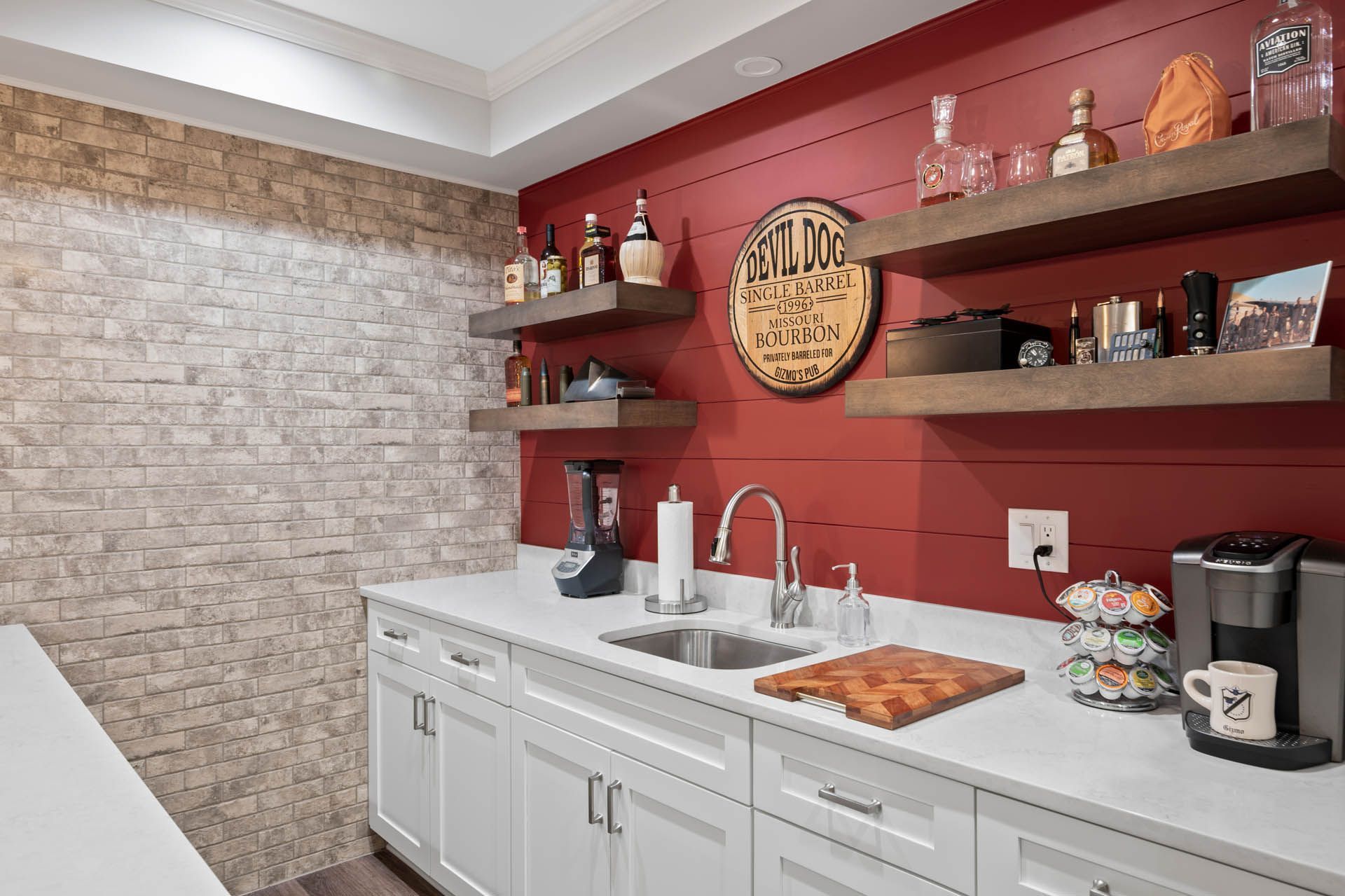 A home bar with a red wall, white cabinets, and stone accent wall. Shelves display liquor and decor above the sink and countertop.