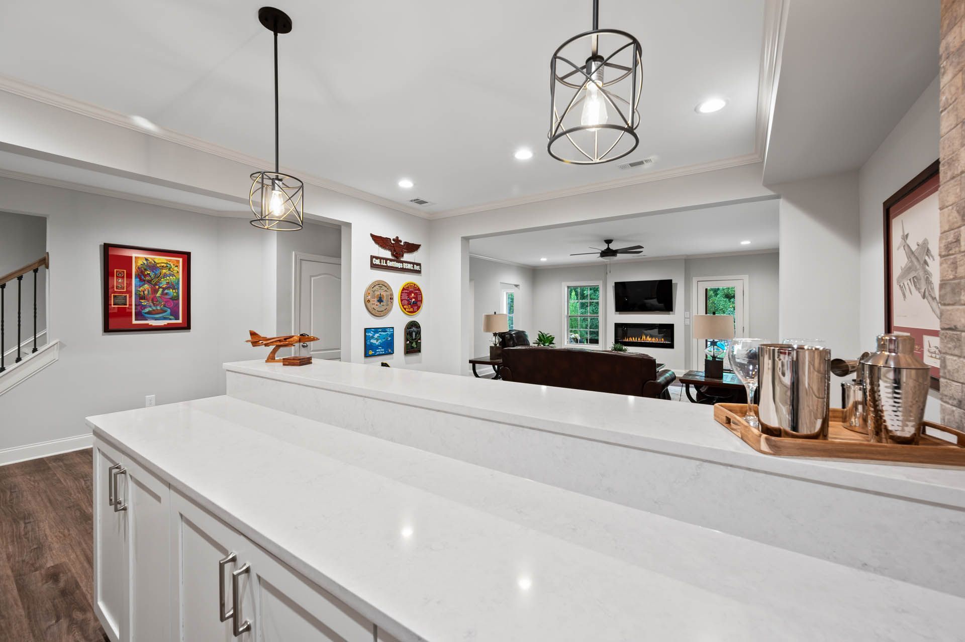 Interior of a basement bar with white countertops, pendant lights, and a view into a living area with a fireplace and TV.