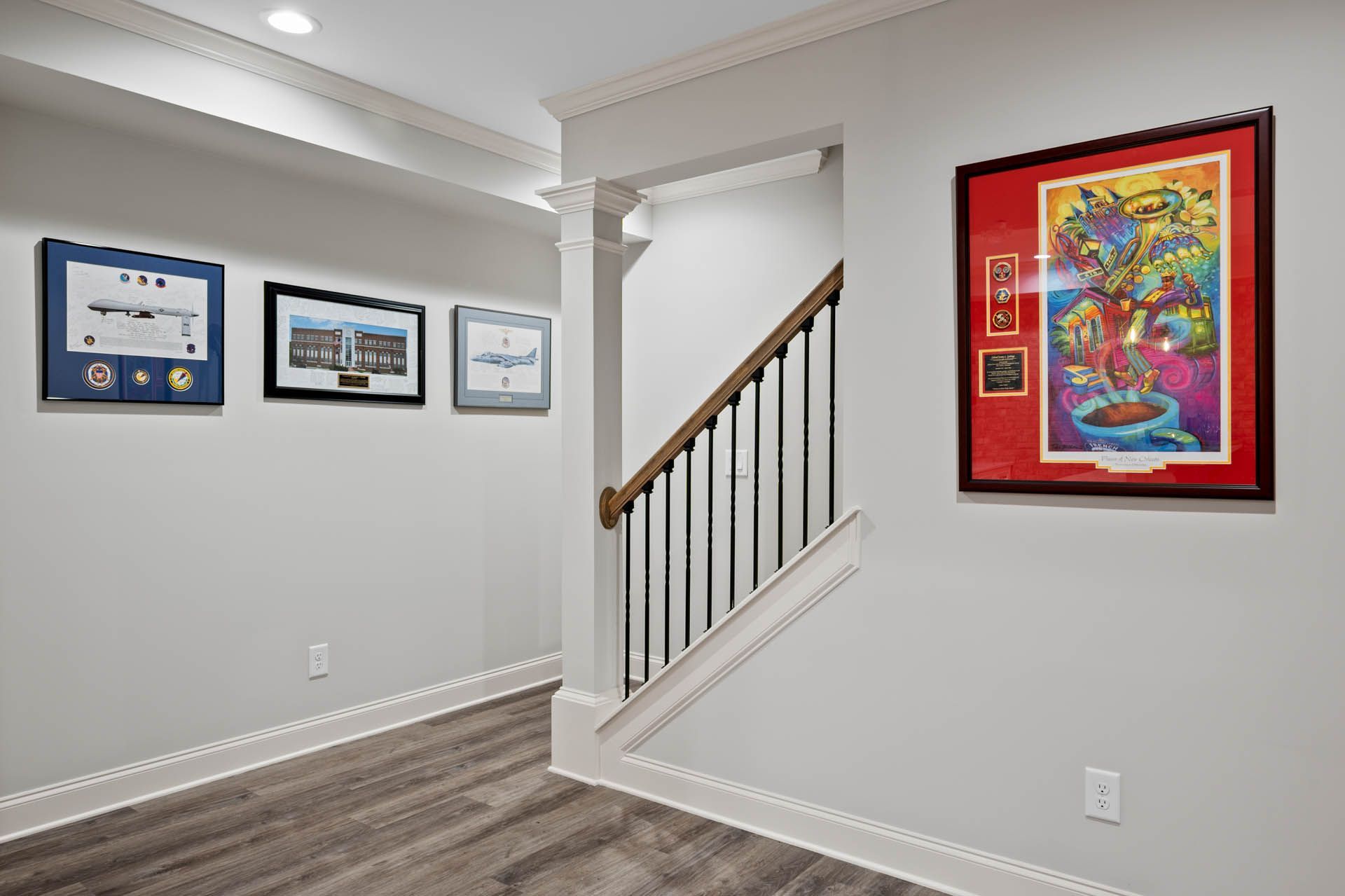Hallway with framed artwork and a staircase. Gray walls, dark wood flooring, and white trim. A colorful print is on the right, with other framed items along the left wall.
