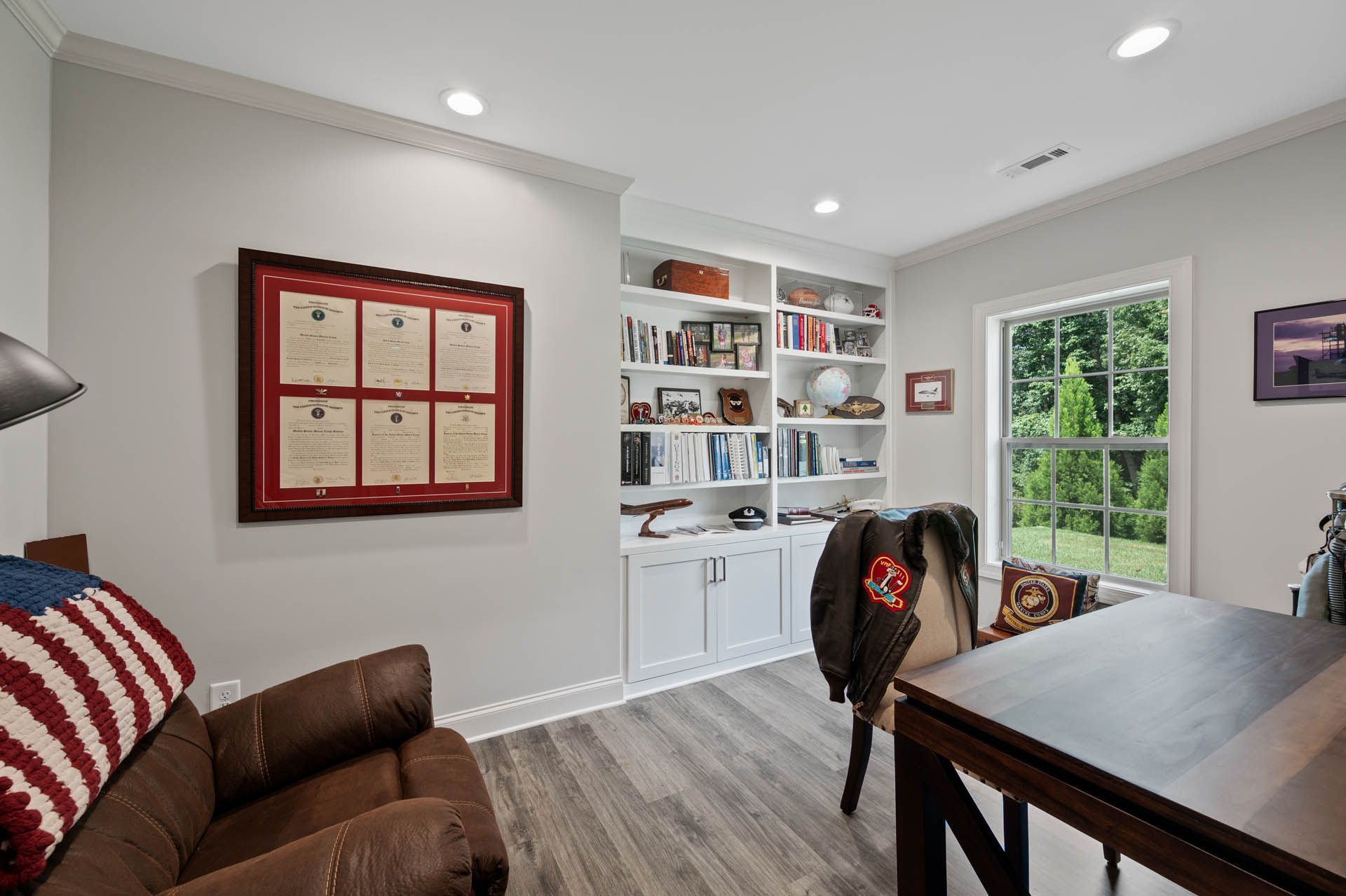 A home office with a brown leather armchair, built-in white bookshelves, and a desk near a window overlooking greenery. A framed document hangs on the wall.