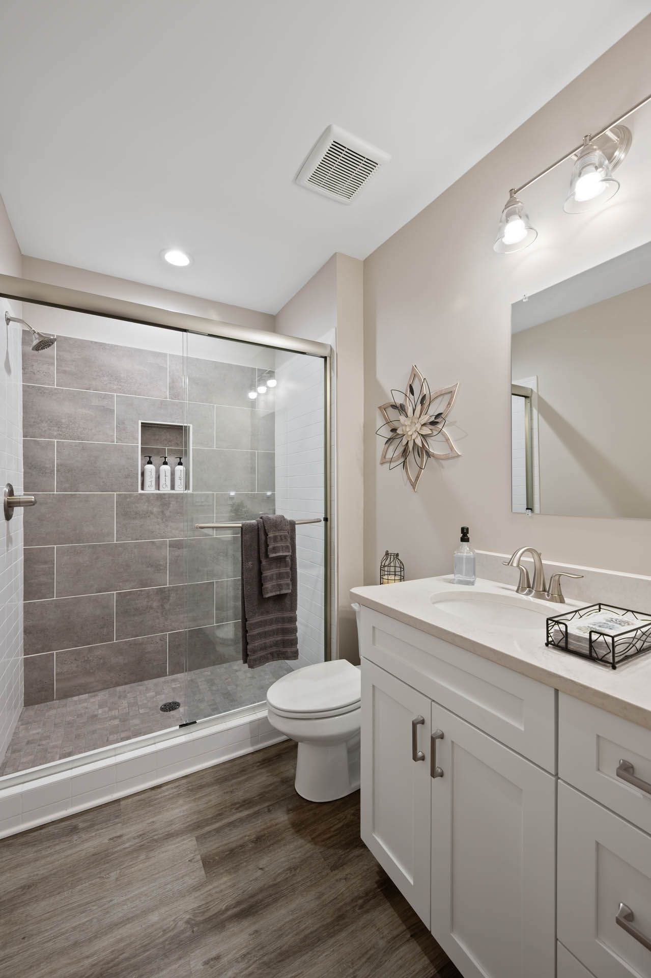 A modern bathroom with a glass shower, white vanity, and gray tile. Wooden floor and neutral walls.