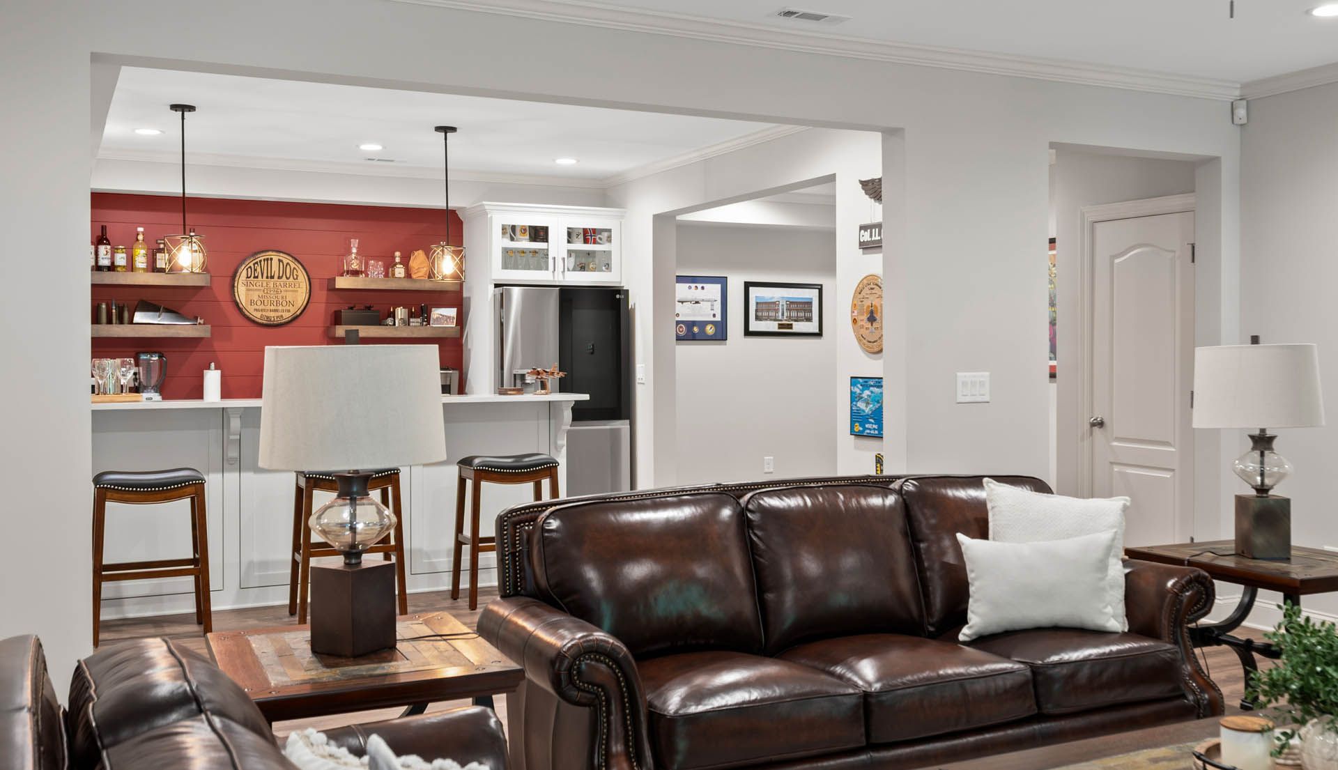 Living room with a brown leather couch, lamp, and rustic coffee table; an opening leads to a kitchen with a red wall and bar.