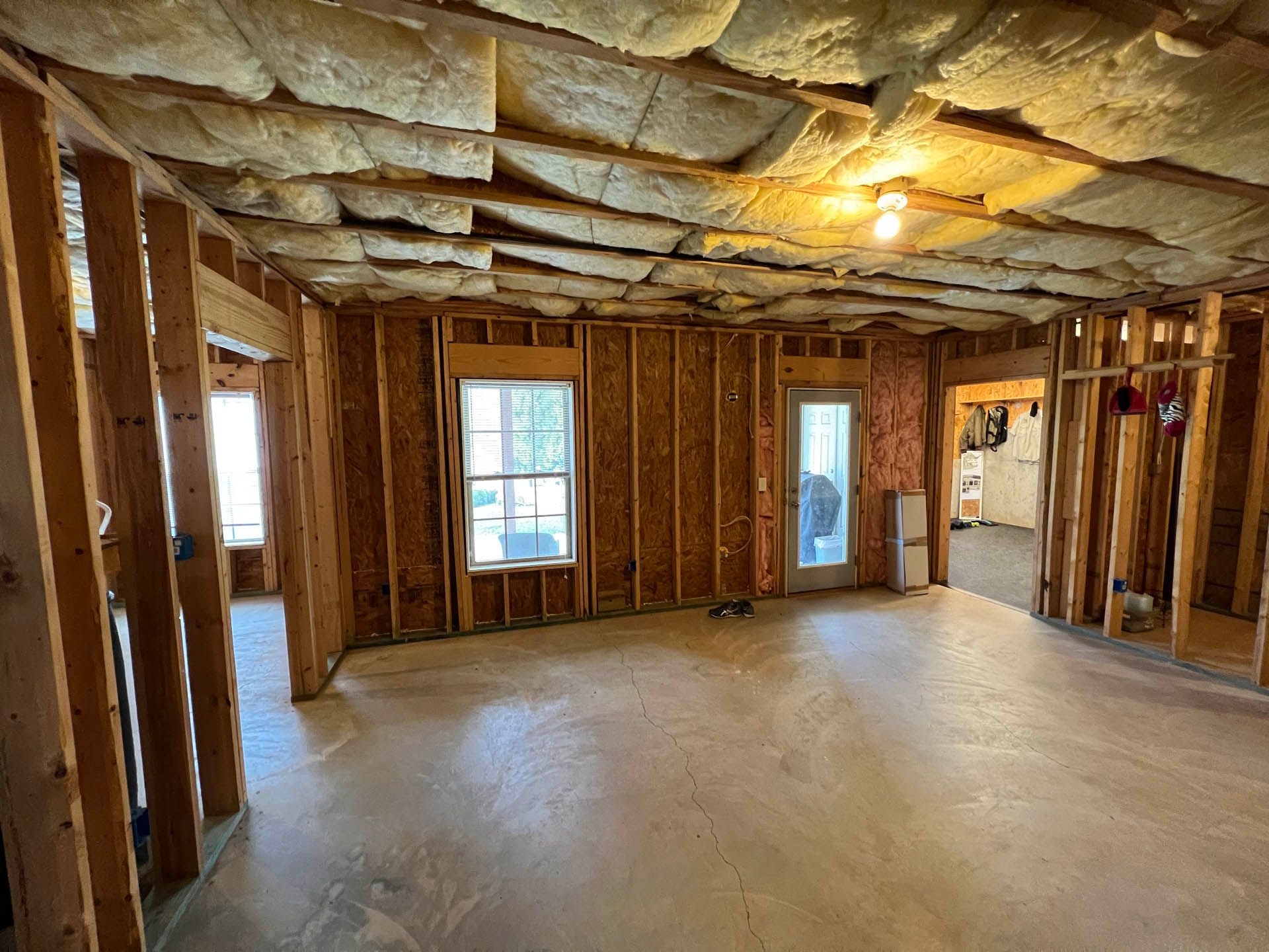 Interior of a room under construction with exposed wooden studs, insulation in the ceiling, and a concrete floor. Two windows and a doorway are visible.