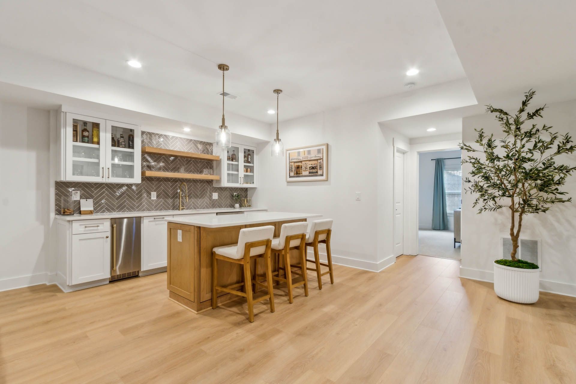 Modern kitchen with a wooden island, white cabinets, and light wood flooring. Bar stools line the island. A doorway leads to another room.