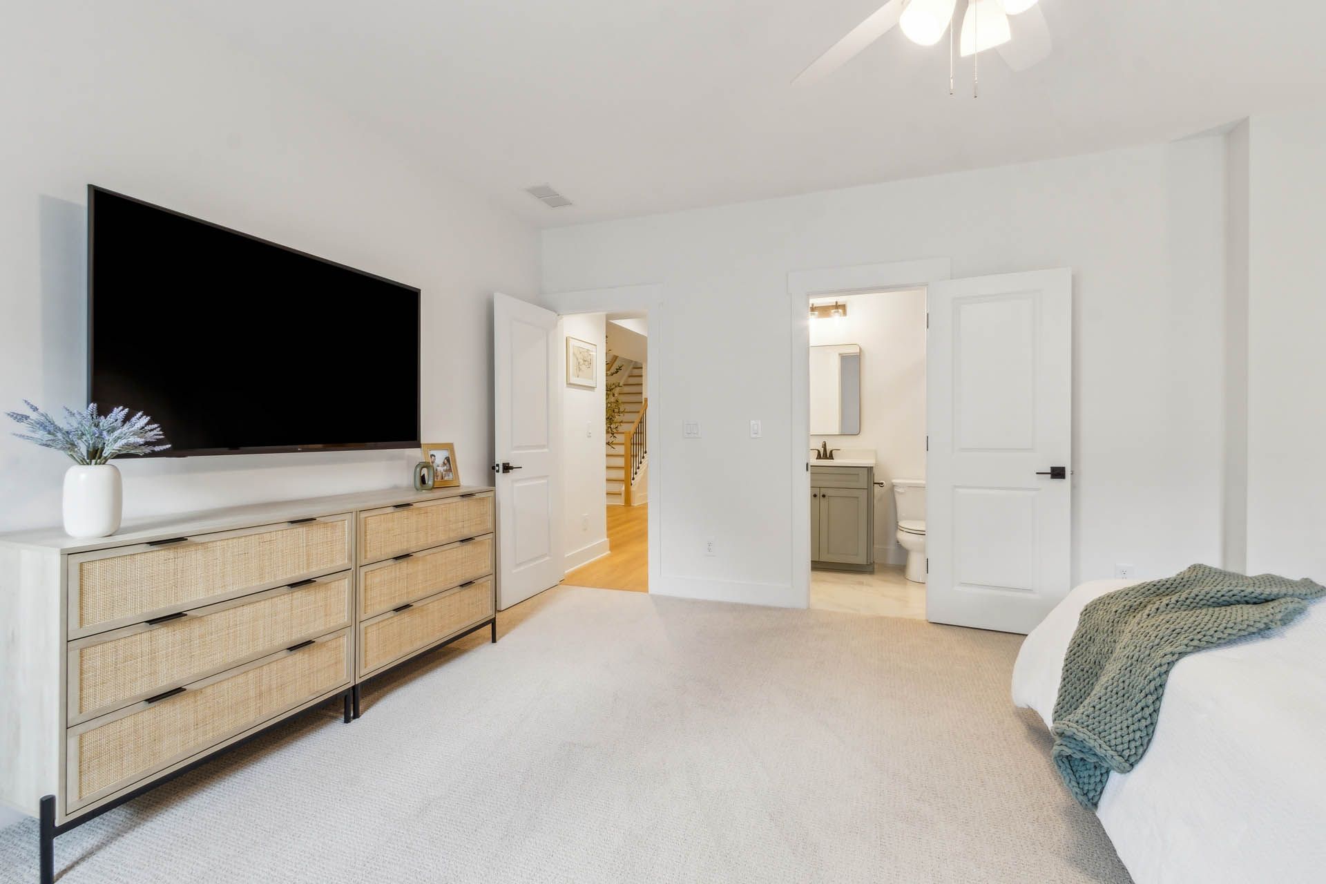 A bedroom with a white rug, dresser with a large TV, and doors leading to a bathroom and hallway.