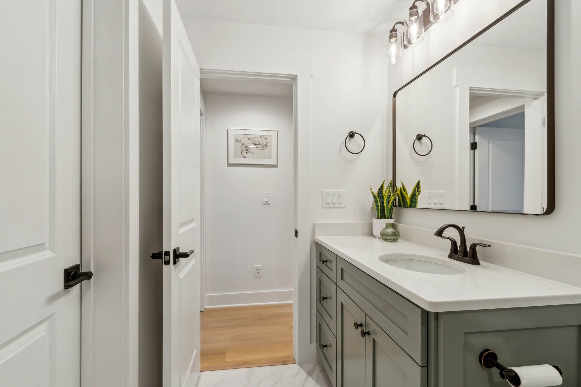 Bathroom with a white vanity, marble countertop, large mirror, and a door leading to another room with wooden flooring.