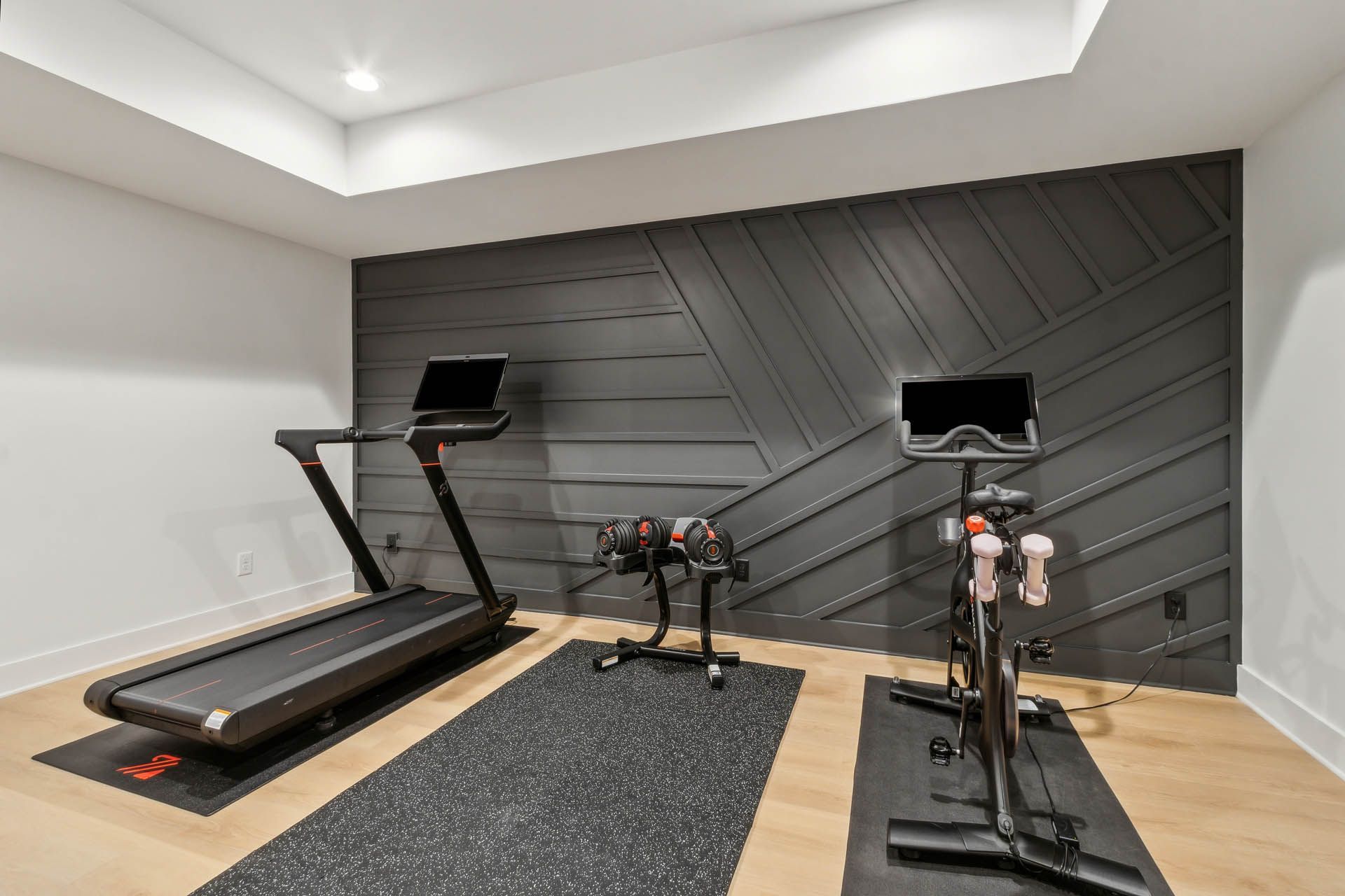 Home gym with a black accent wall. Treadmill, spin bike, and weights on mats in a white-walled room.