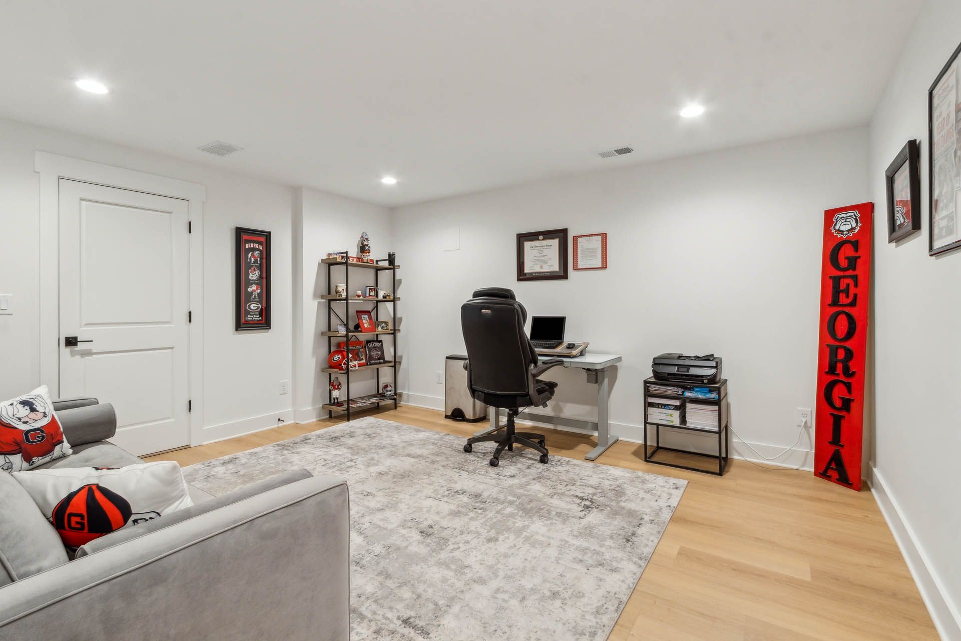 Home office featuring a desk with a computer, a gray sofa, and a bookshelf with University of Georgia decor.