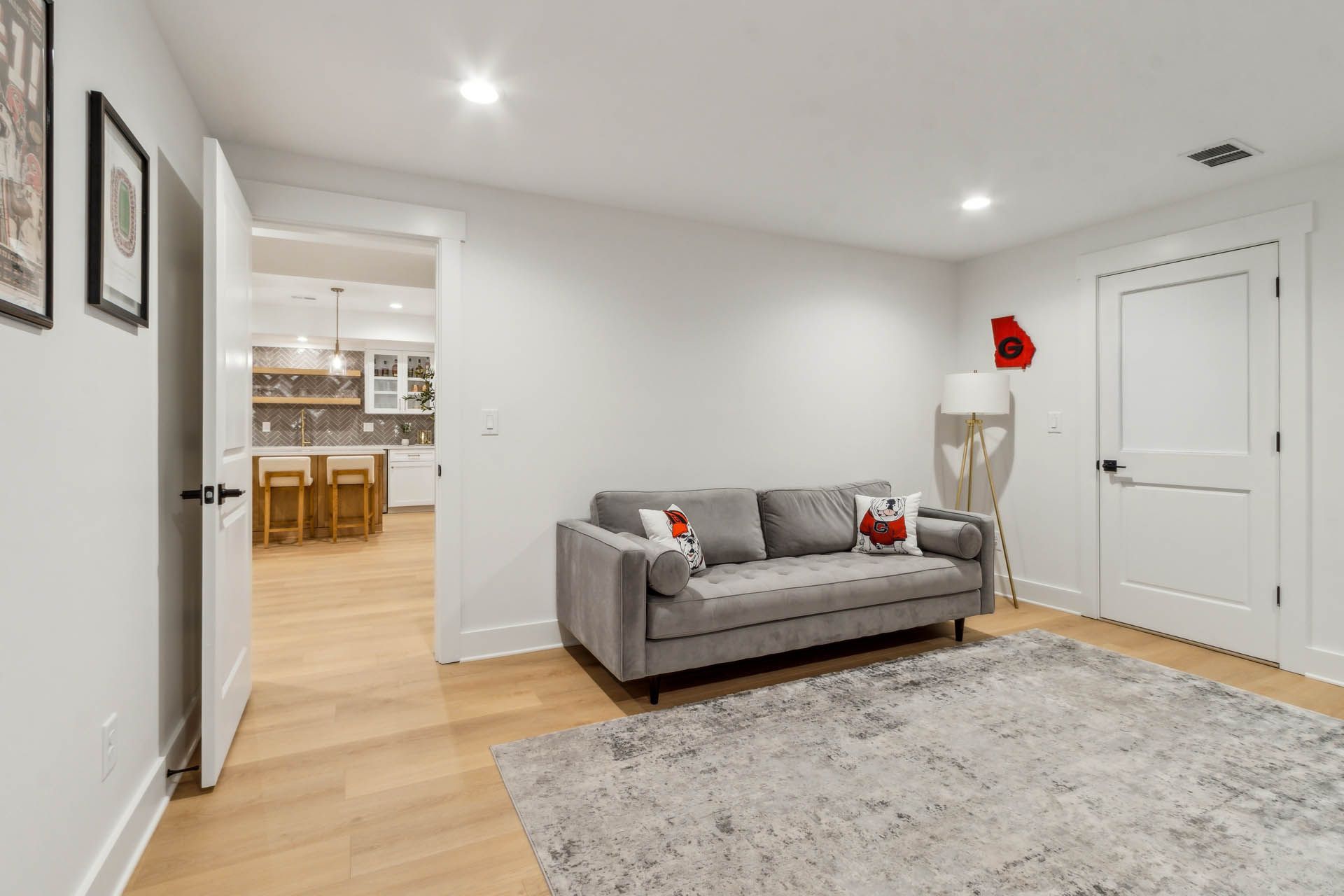 A modern living room with a gray couch, rug, and white walls. An open doorway reveals a kitchen.