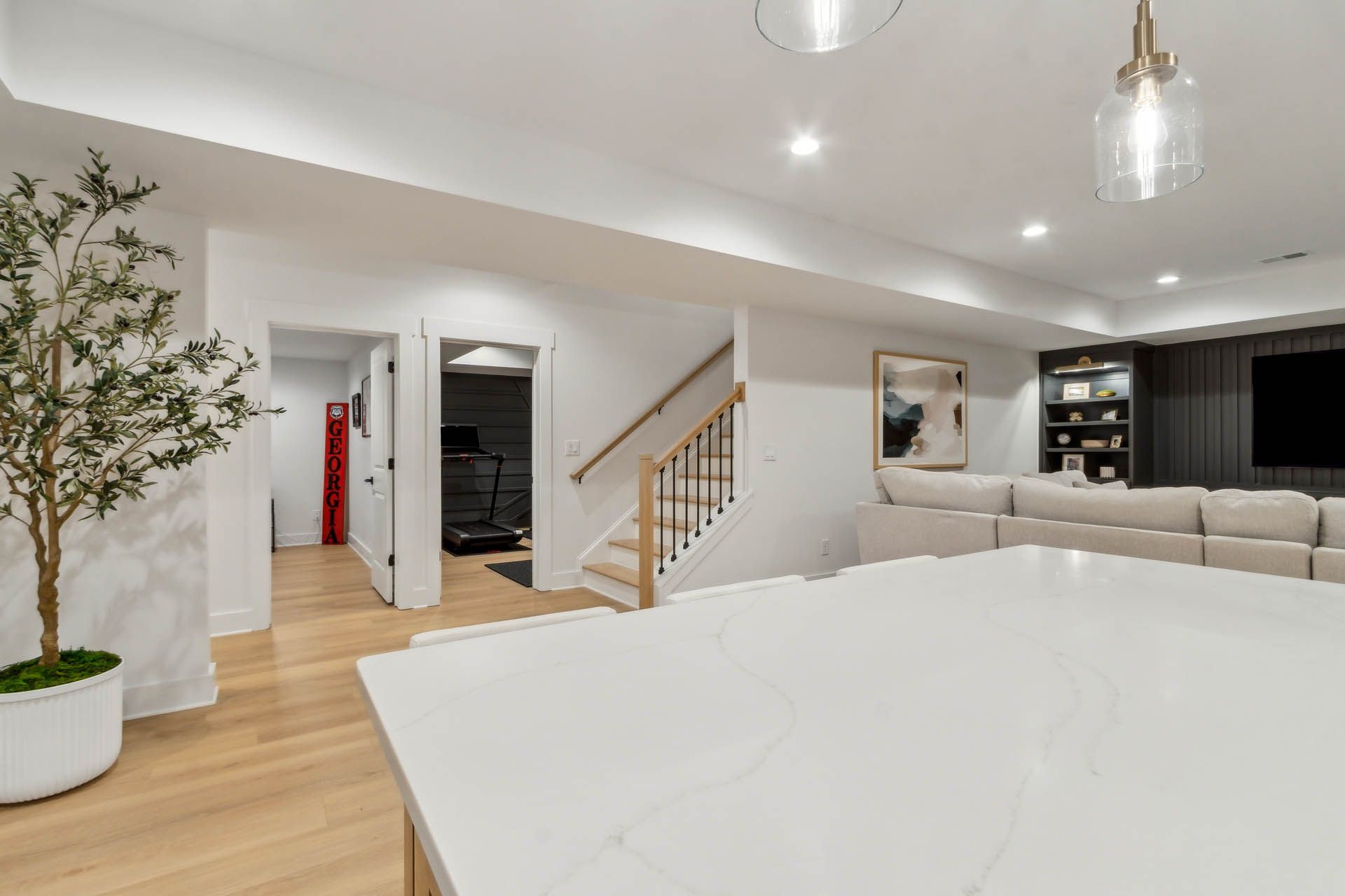 Spacious, modern basement interior featuring a kitchen island, stairs, seating area, and doorway to another room with a treadmill.