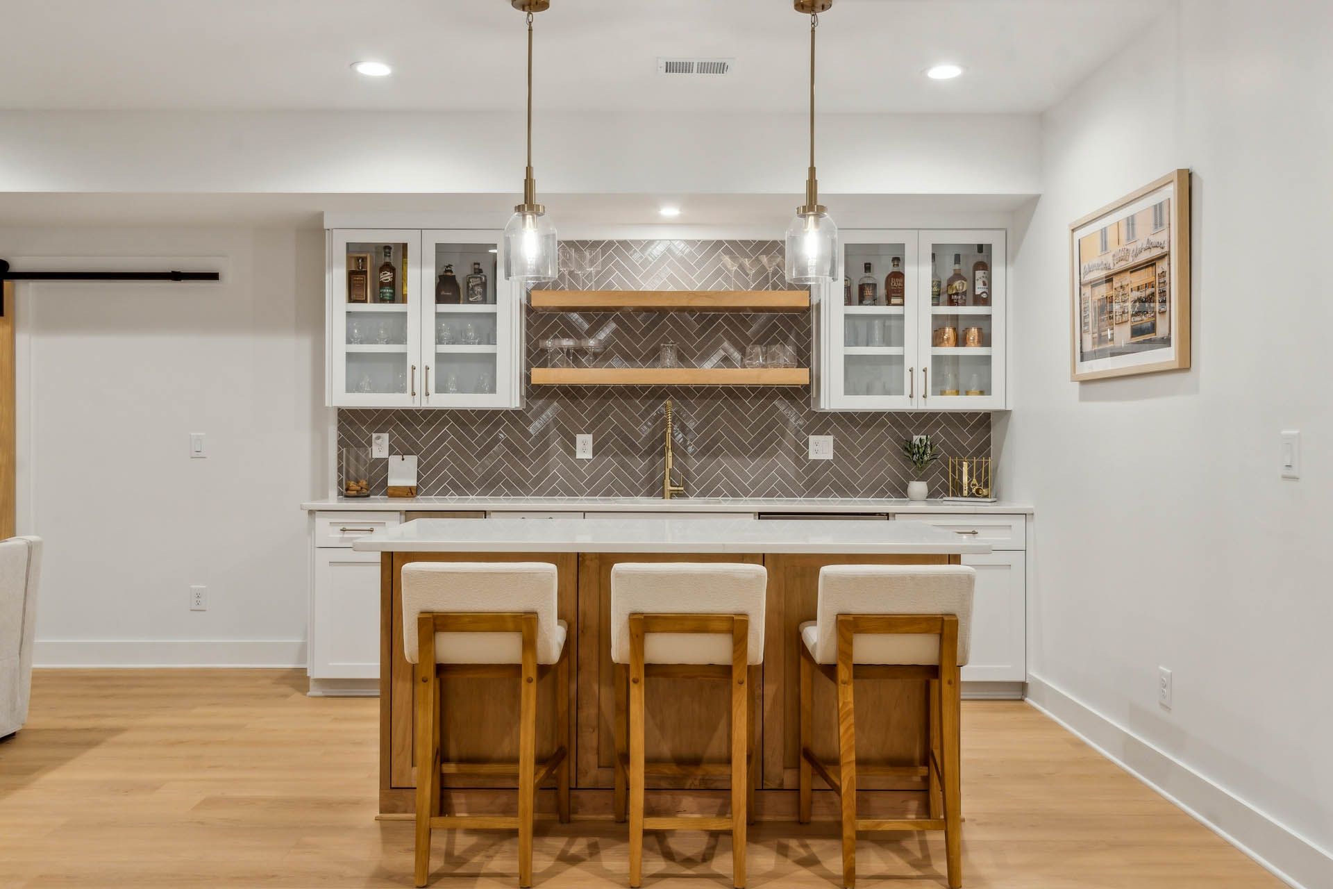 A home bar with a white countertop, wooden cabinets, and three bar stools. It has a neutral color palette, with light wood flooring.