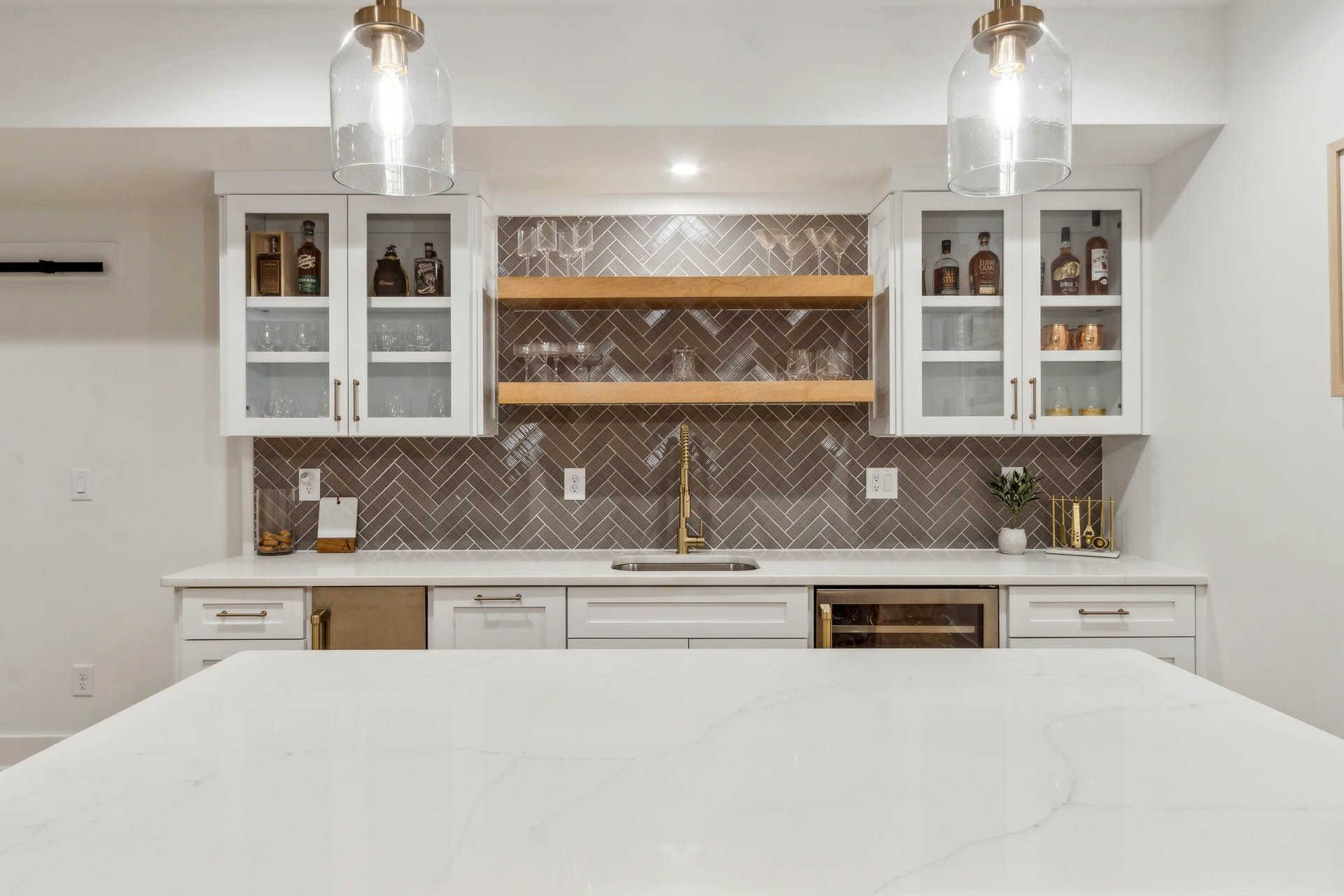 White bar area with a gray herringbone tile backsplash, open shelving, and glass-front cabinets. Overhead, two clear pendant lights hang.