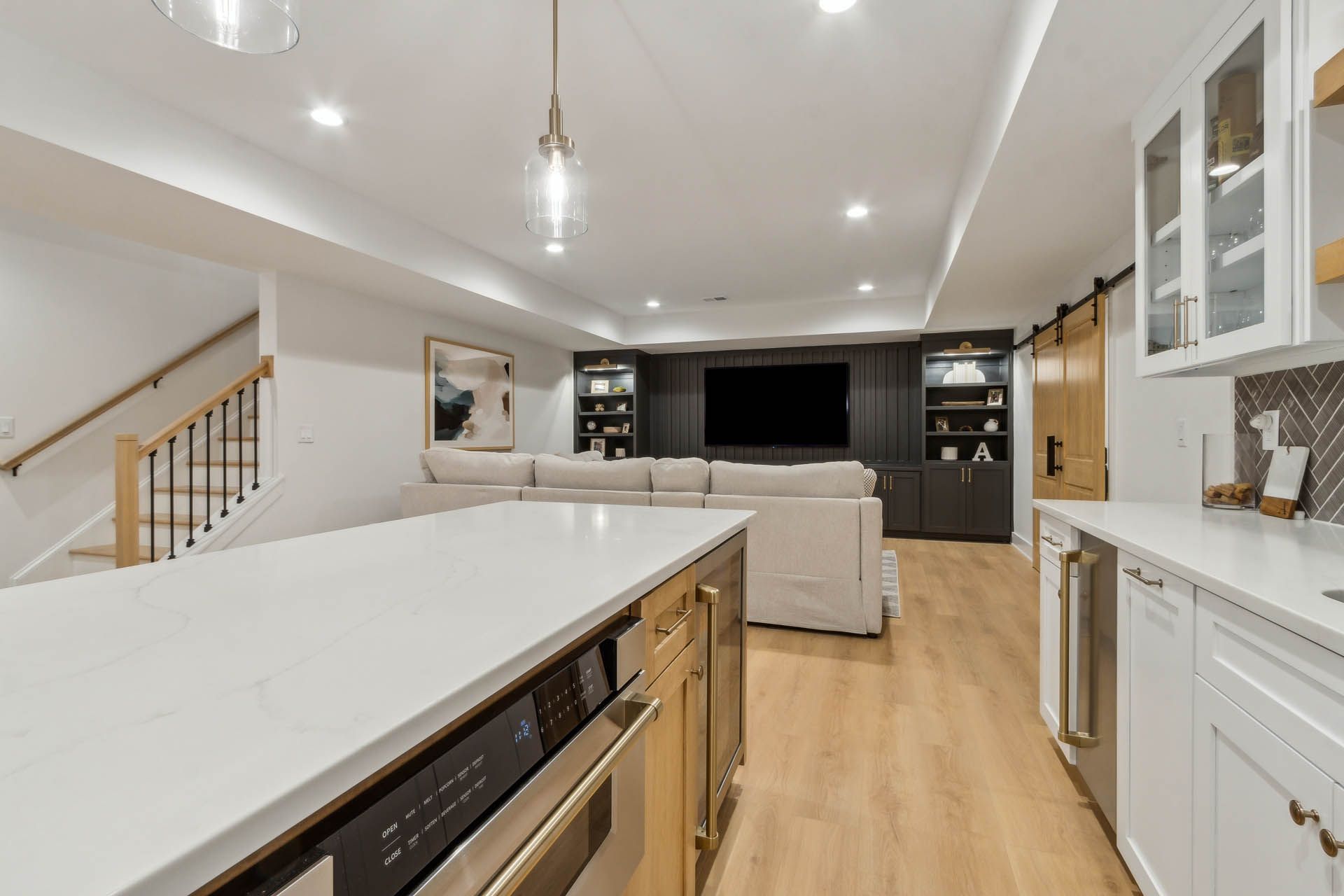 Modern finished basement with a kitchen, living area, and staircase. Features white countertops, a dark entertainment center, and light-colored wood flooring.