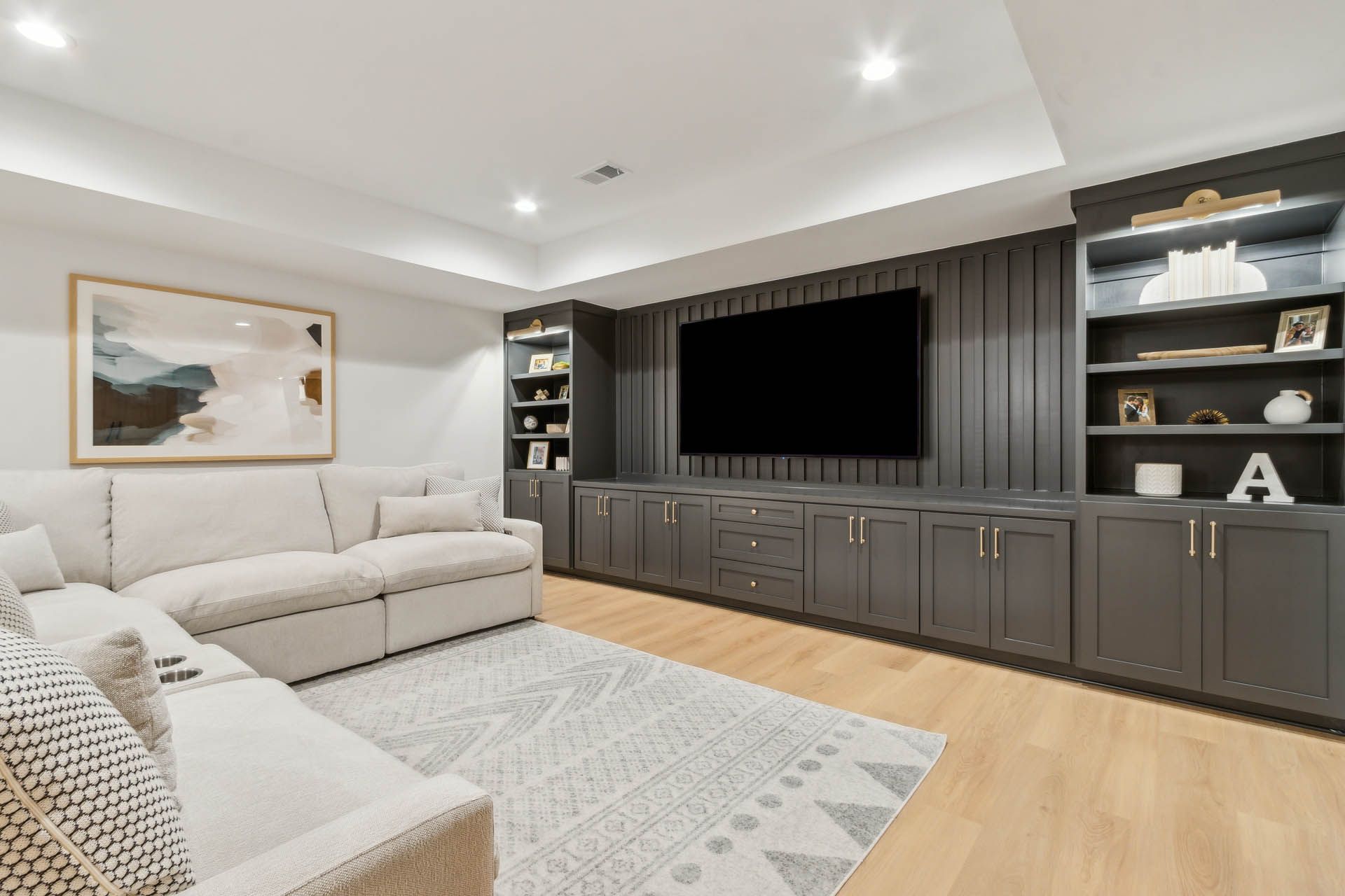 A modern living room with a gray entertainment center housing a large TV.  A white sectional sofa sits opposite a patterned rug.