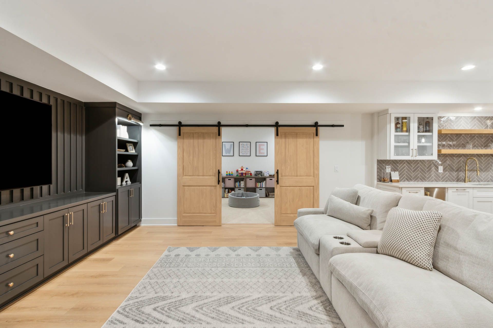 Modern basement living space with wood flooring, a large sectional sofa, and dark gray built-in entertainment center. Sliding barn doors lead to another room.