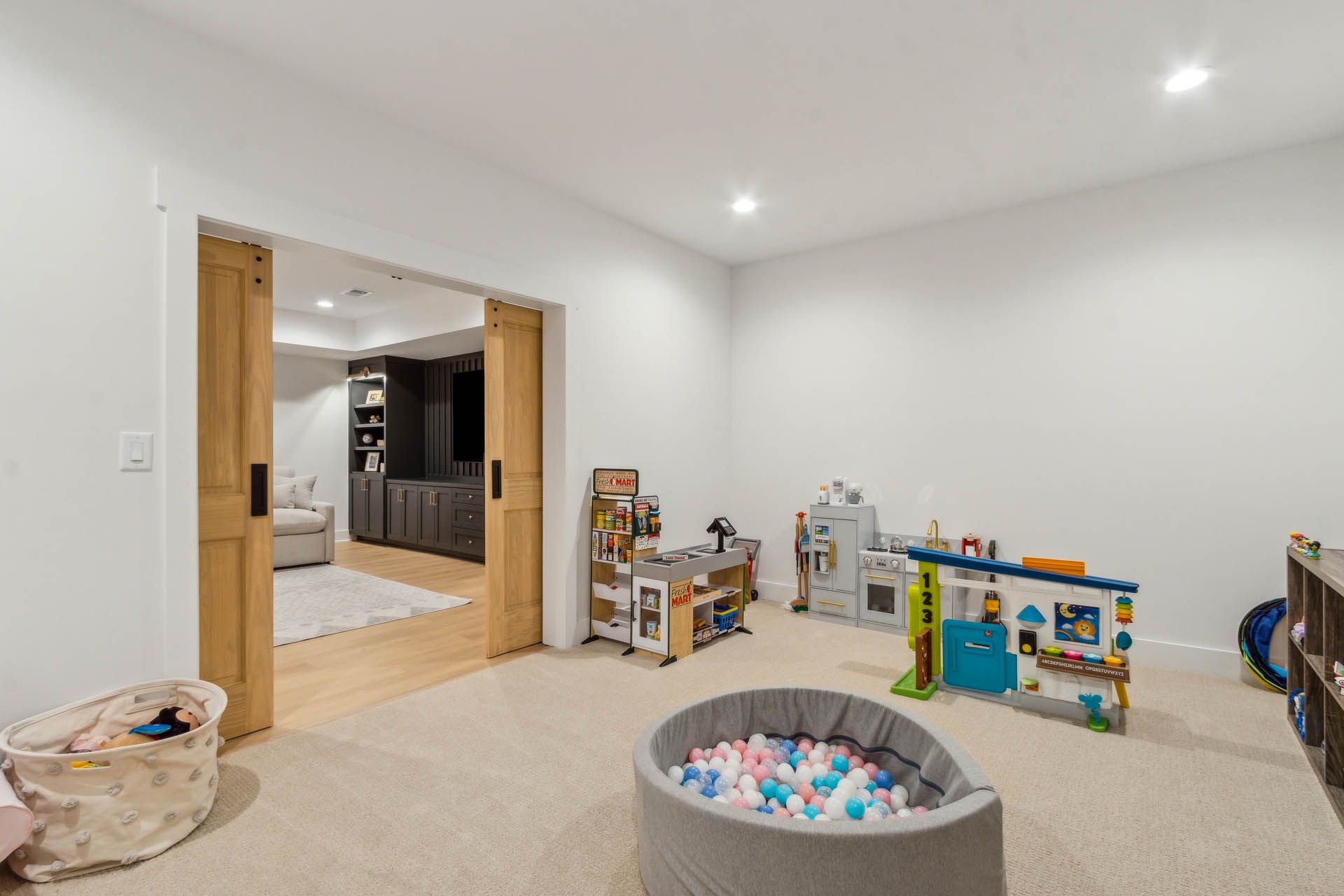 Spacious playroom with light-colored carpet, toys, and a ball pit. Two wooden sliding doors open into another room.