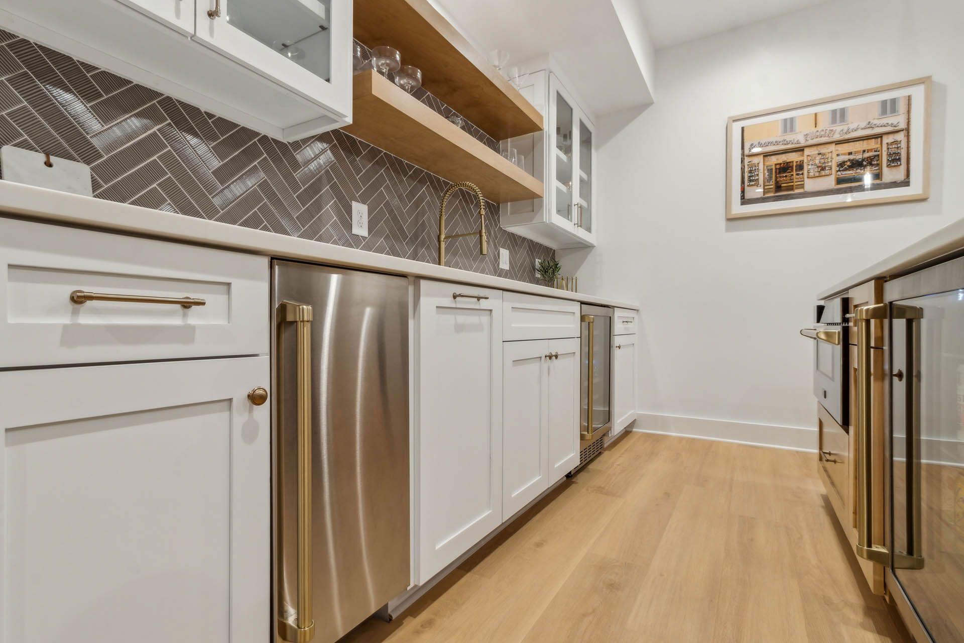 A modern, white kitchenette with stainless steel appliances, light wood shelving, and a gray herringbone backsplash.