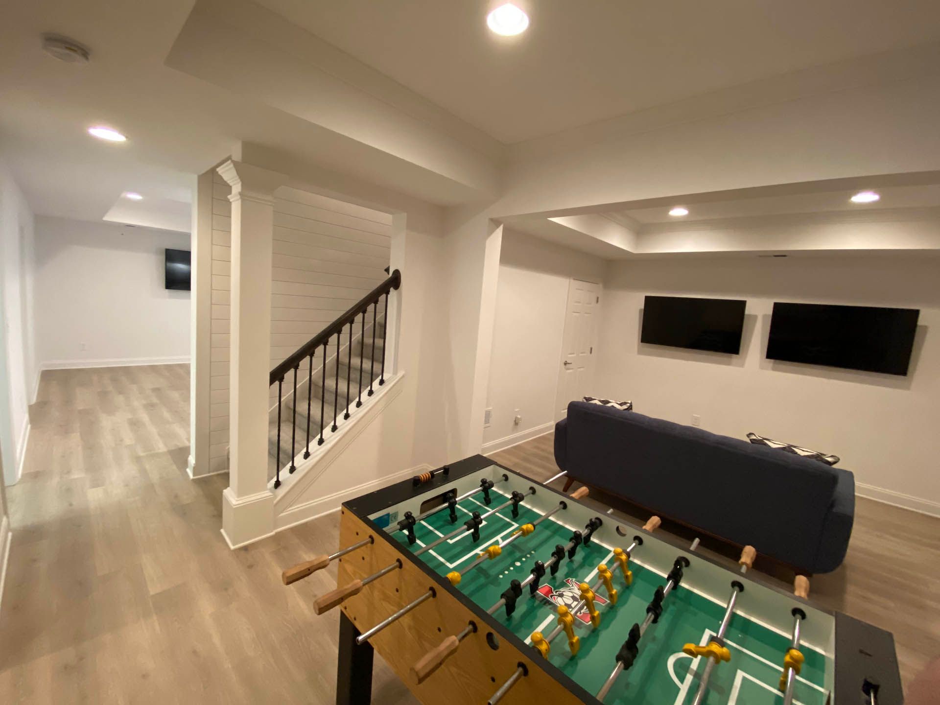 A finished basement featuring a foosball table, sofa, two TVs, and a staircase leading to another room.