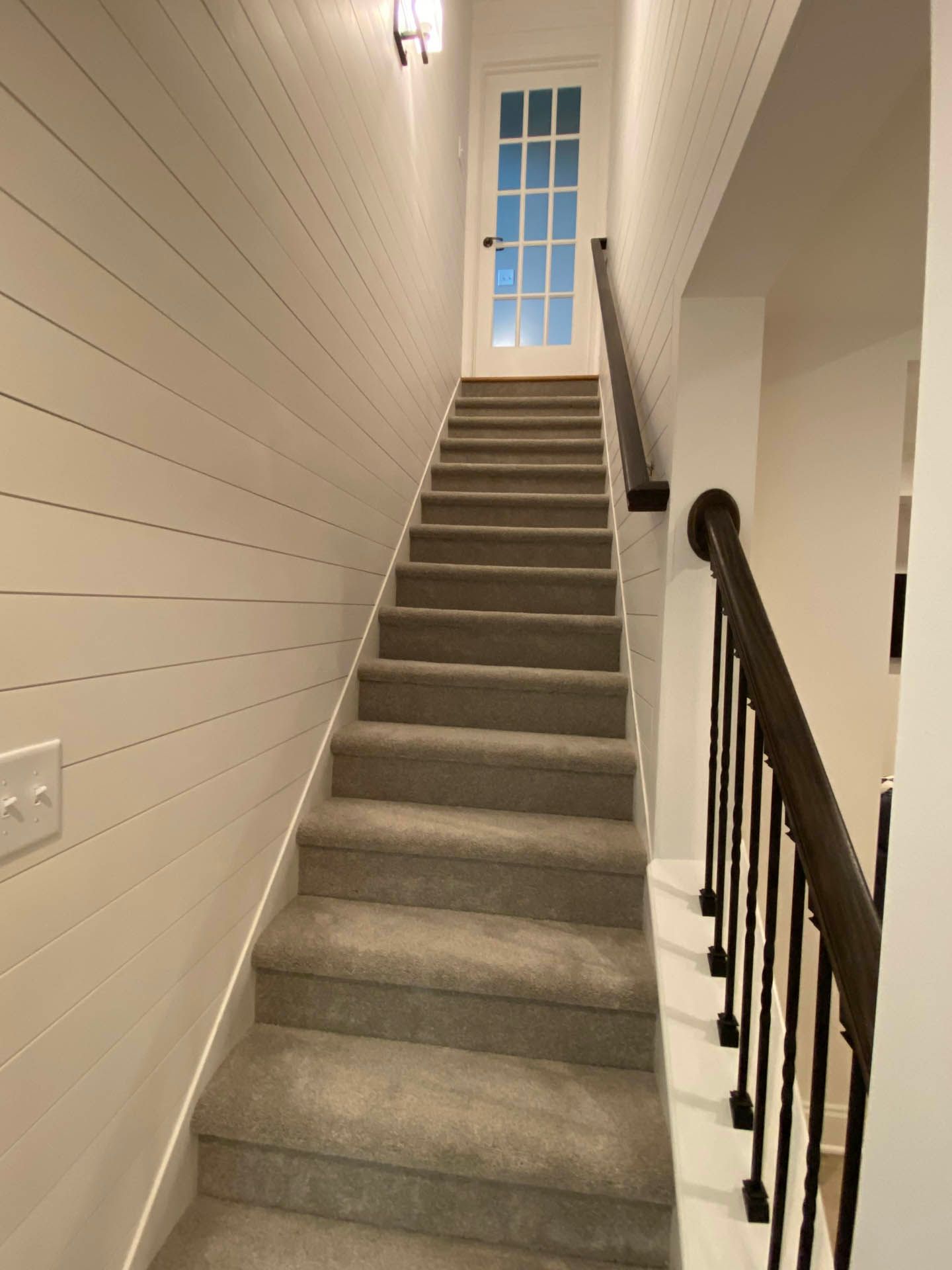 Staircase with carpeted steps leading upwards to a white door. One wall is paneled, and a dark railing runs alongside the stairs.
