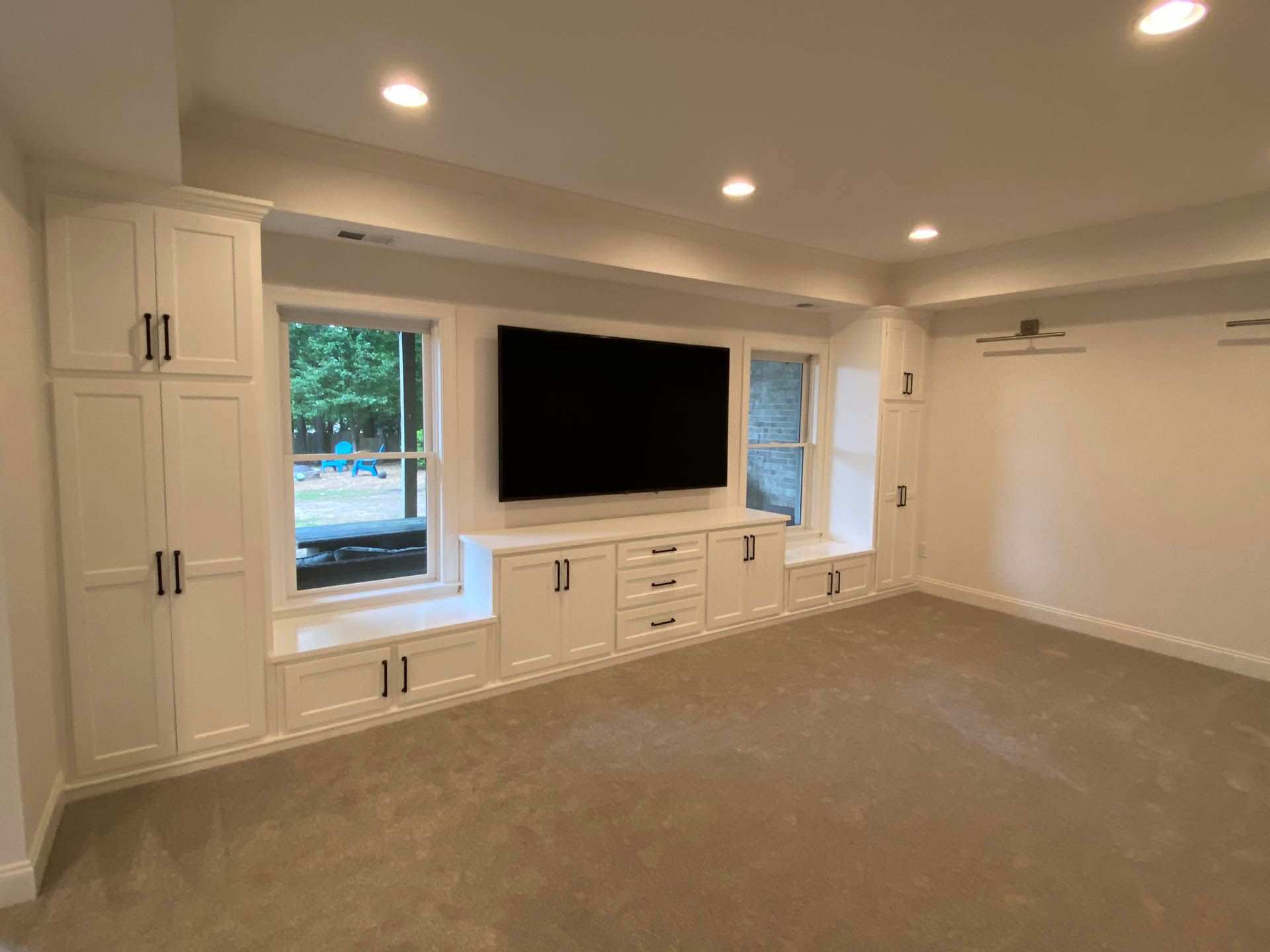 A white built-in entertainment center in a room with a television, cabinets, drawers, and windows; room has grey carpet.