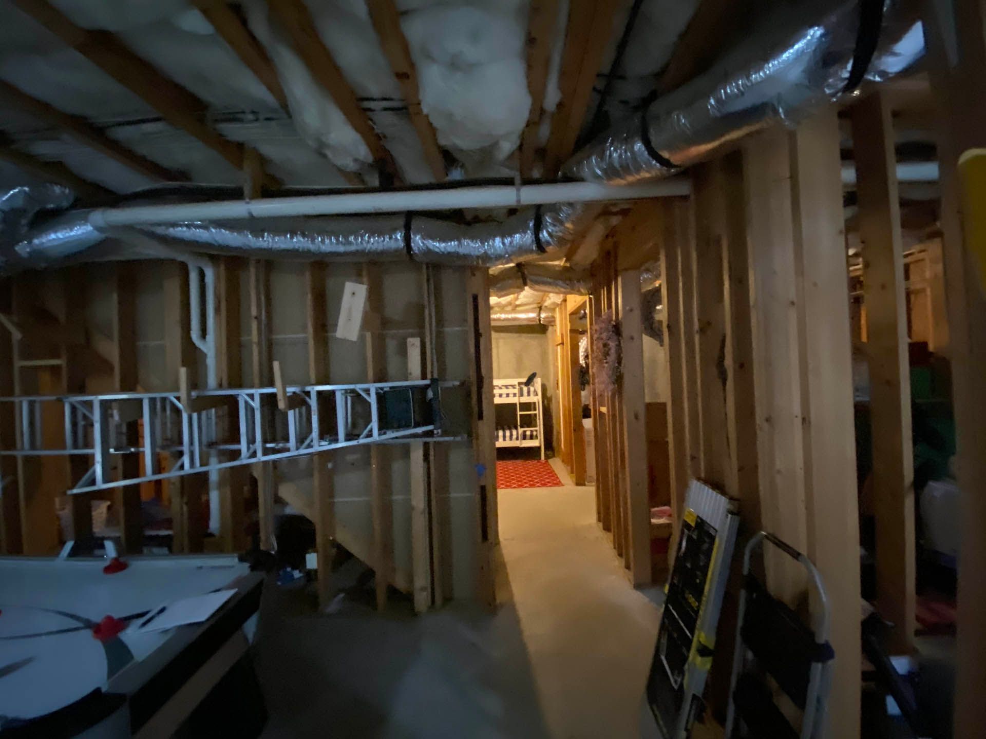 Basement hallway under construction. Exposed wooden studs and ductwork run along the walls and ceiling. A white ladder leans against the wall.