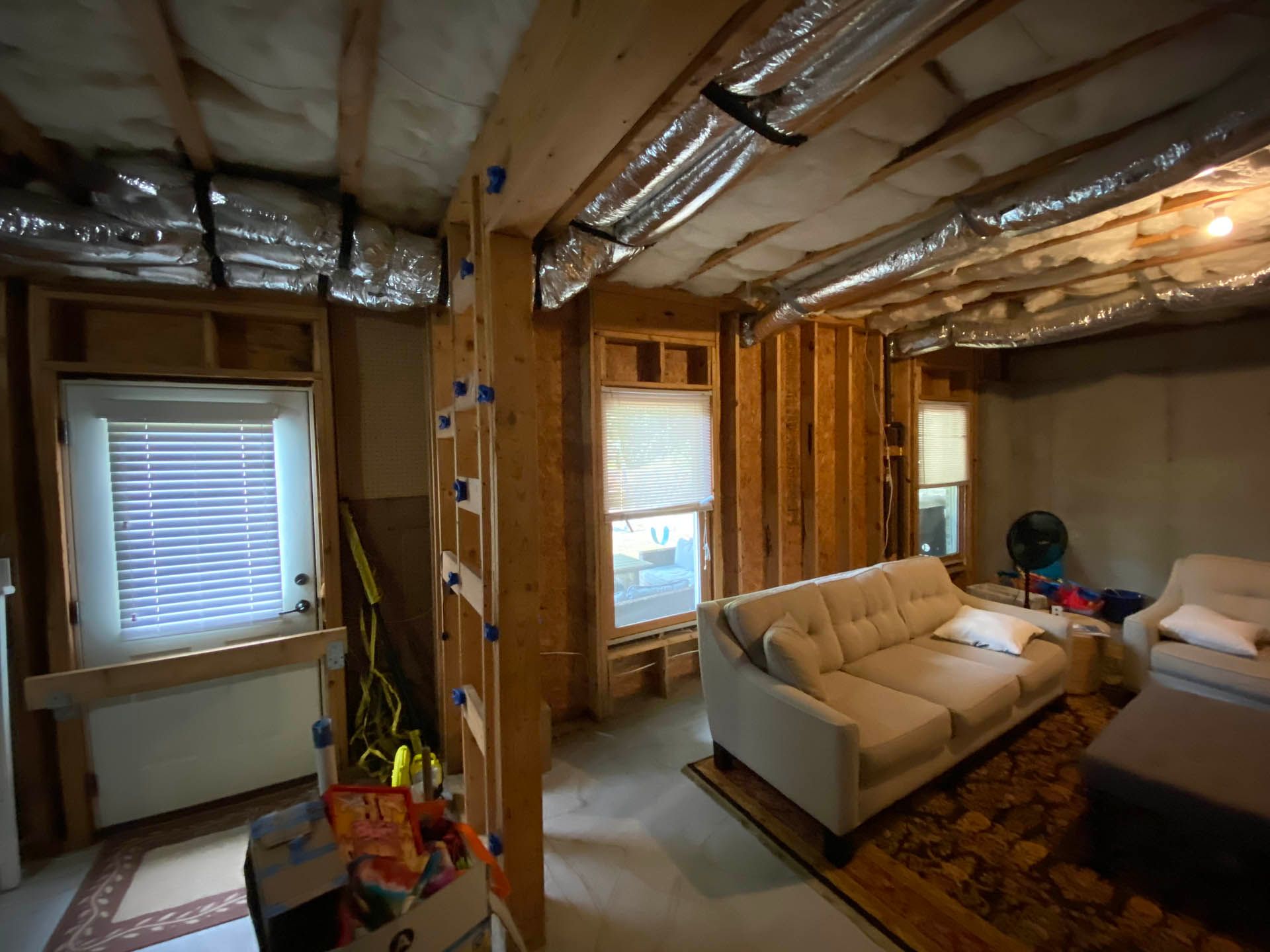 Interior view of an unfinished basement, with exposed wood framing, a couch, windows with blinds, and ductwork.