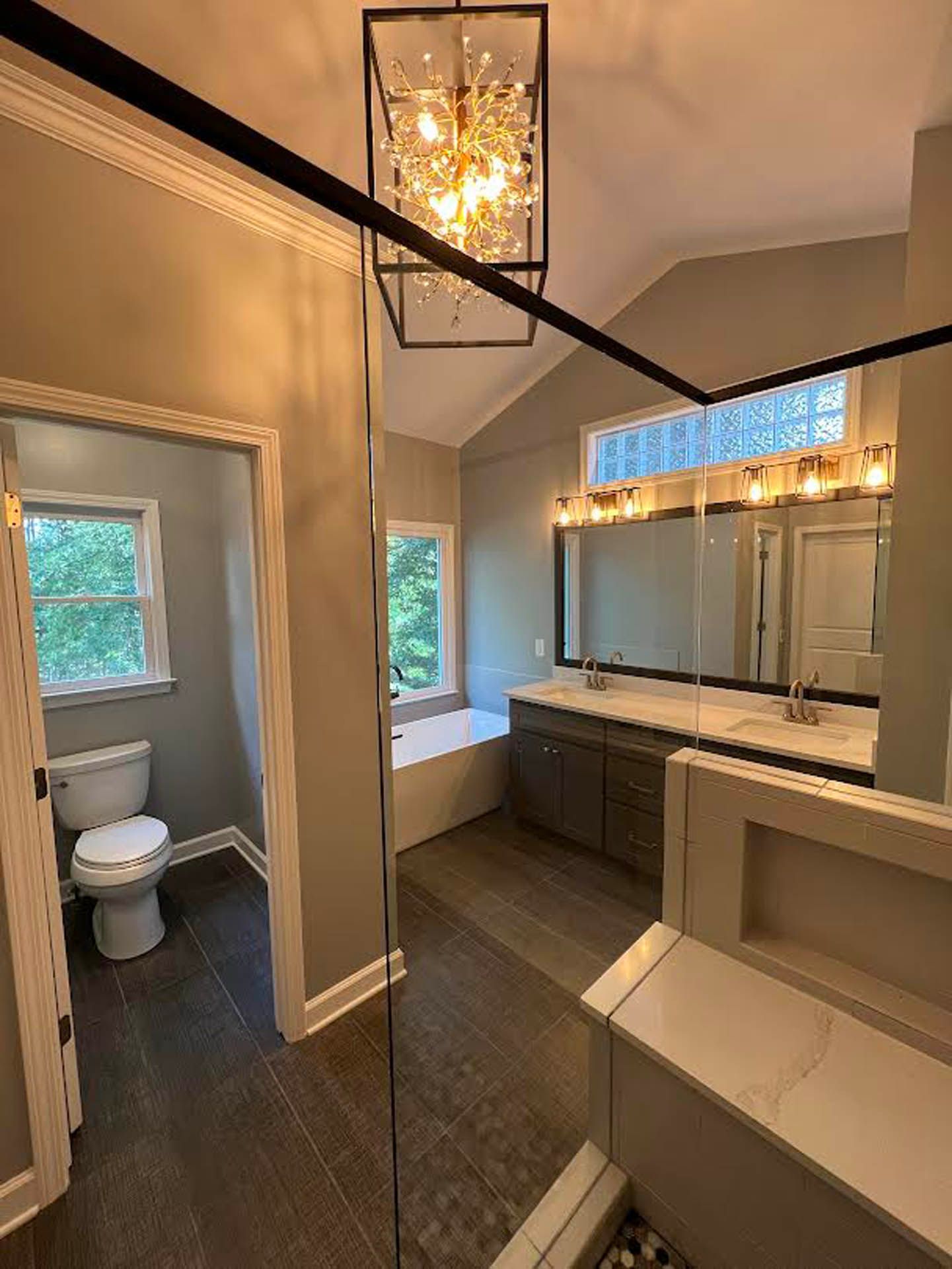 Spacious bathroom with a toilet, double vanity, and a bathtub. Brown floors, light gray walls, and a black light fixture.