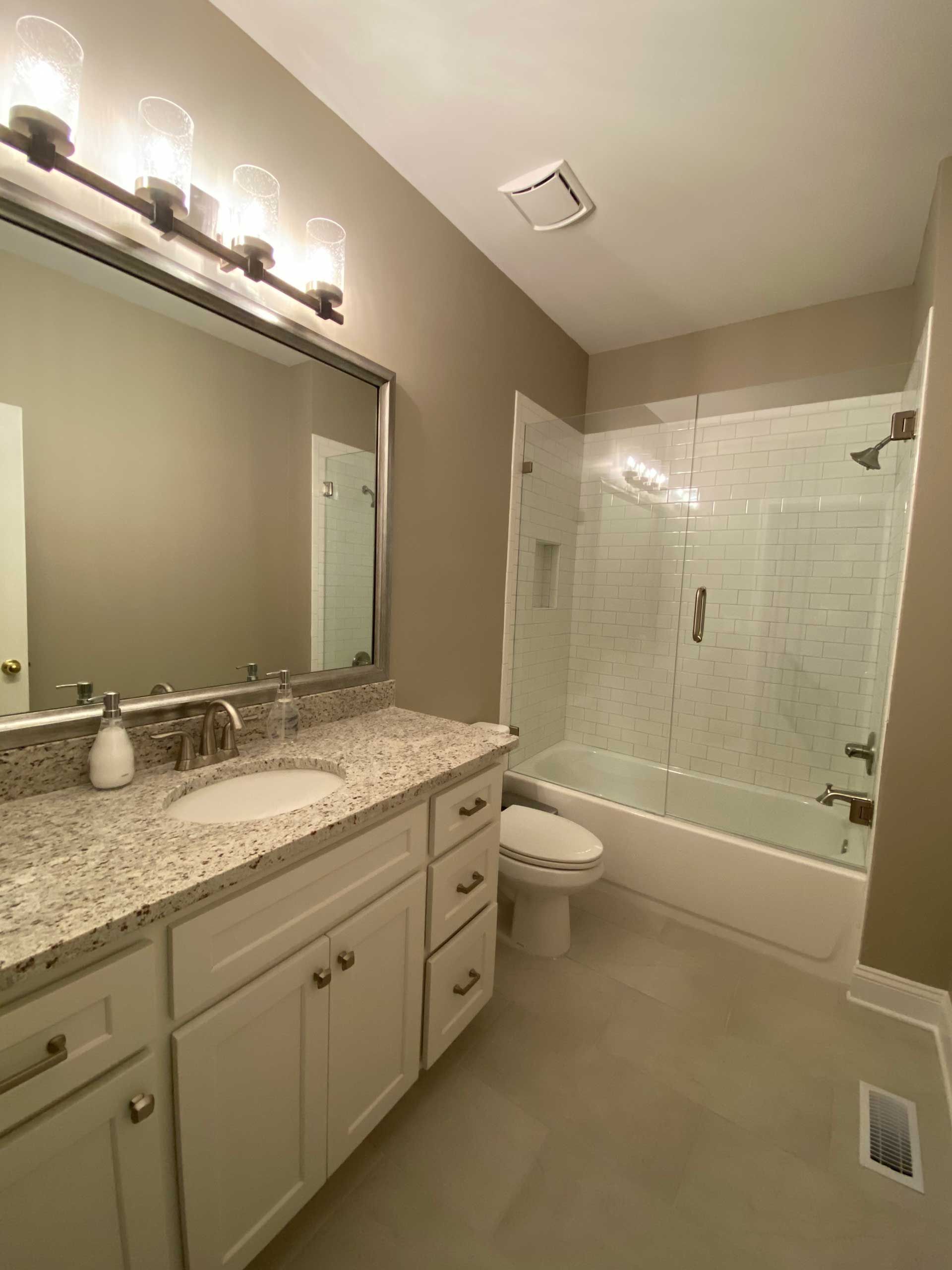 Bathroom with white vanity, granite countertop, large mirror, white toilet, and tiled shower with a glass door. Beige walls and gray tile flooring.