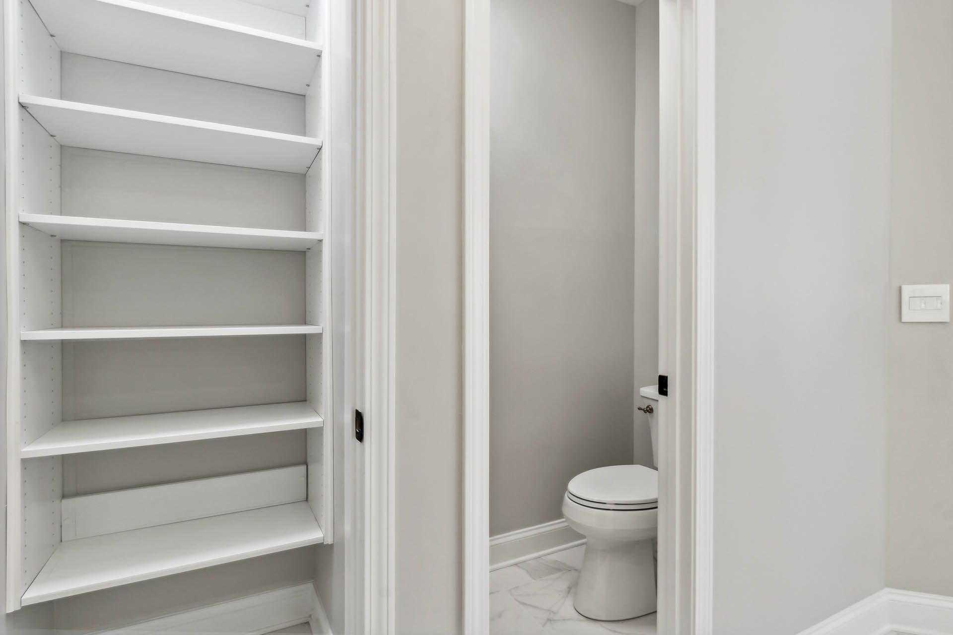 A narrow hallway leads to a toilet, with white shelves visible on the left. The walls are gray and the toilet is white.