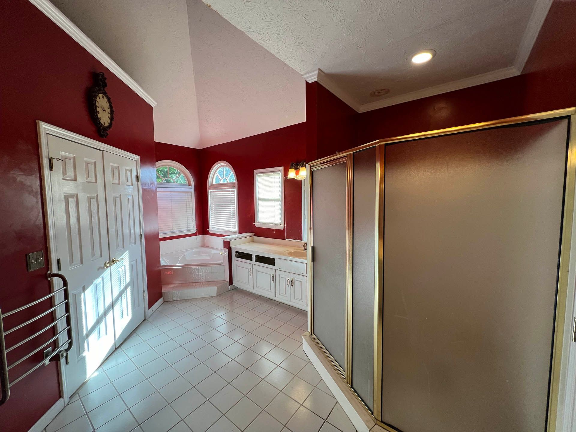 Spacious bathroom with red walls, white tile floor, and a large glass shower. Features a soaking tub, white vanity, and arched windows.