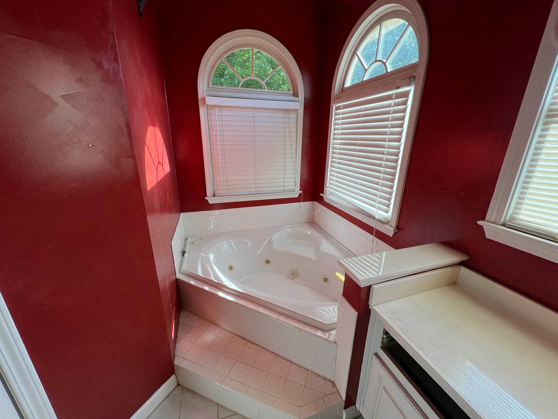 Red bathroom with a jacuzzi tub under arched windows with blinds. White countertop and cabinets are on the right.