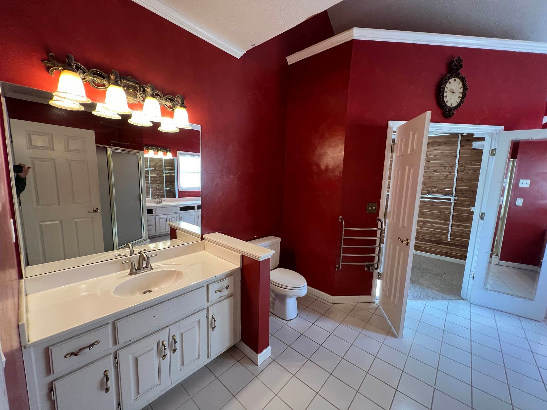 A bathroom with a white vanity, toilet, and floor against red walls. A clock hangs above a doorway to a shower with bamboo-style doors.