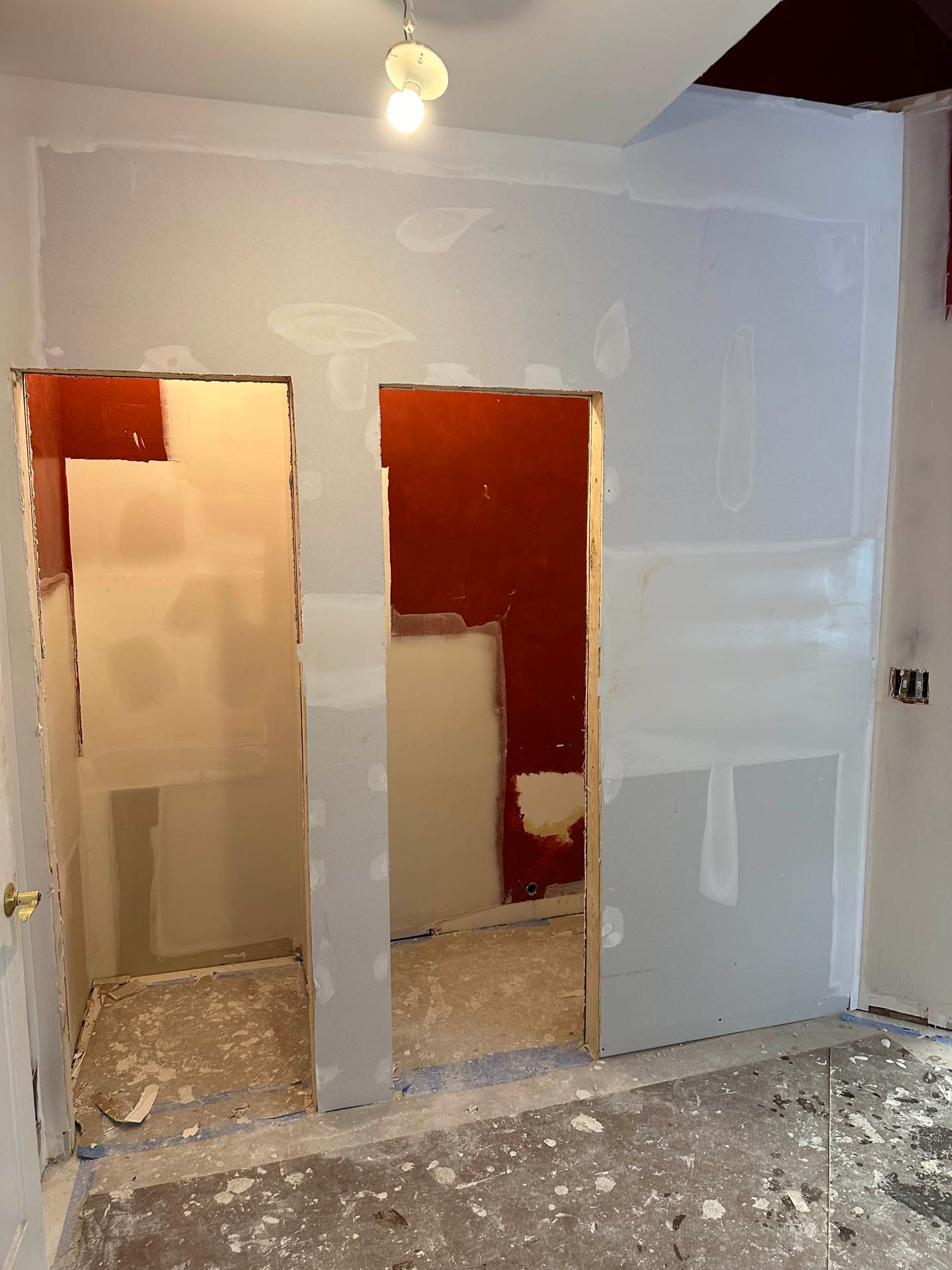Room under construction with two unfinished doorways, drywall, and red painted sections. Light-colored walls, debris on the floor, and a ceiling light are visible.