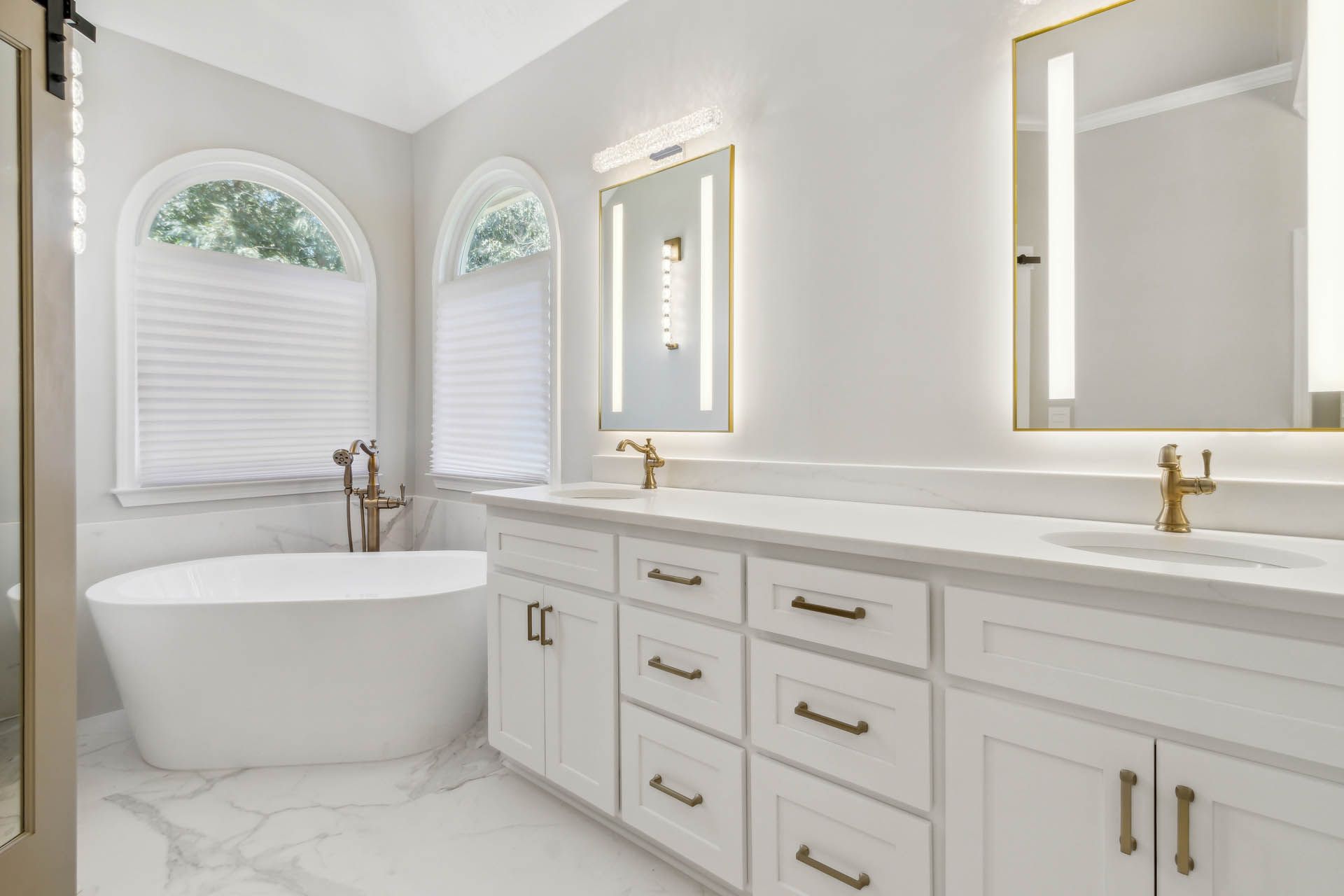 A bright white bathroom with a soaking tub, double vanity, and arched windows. Gold fixtures and hardware accent the clean lines.