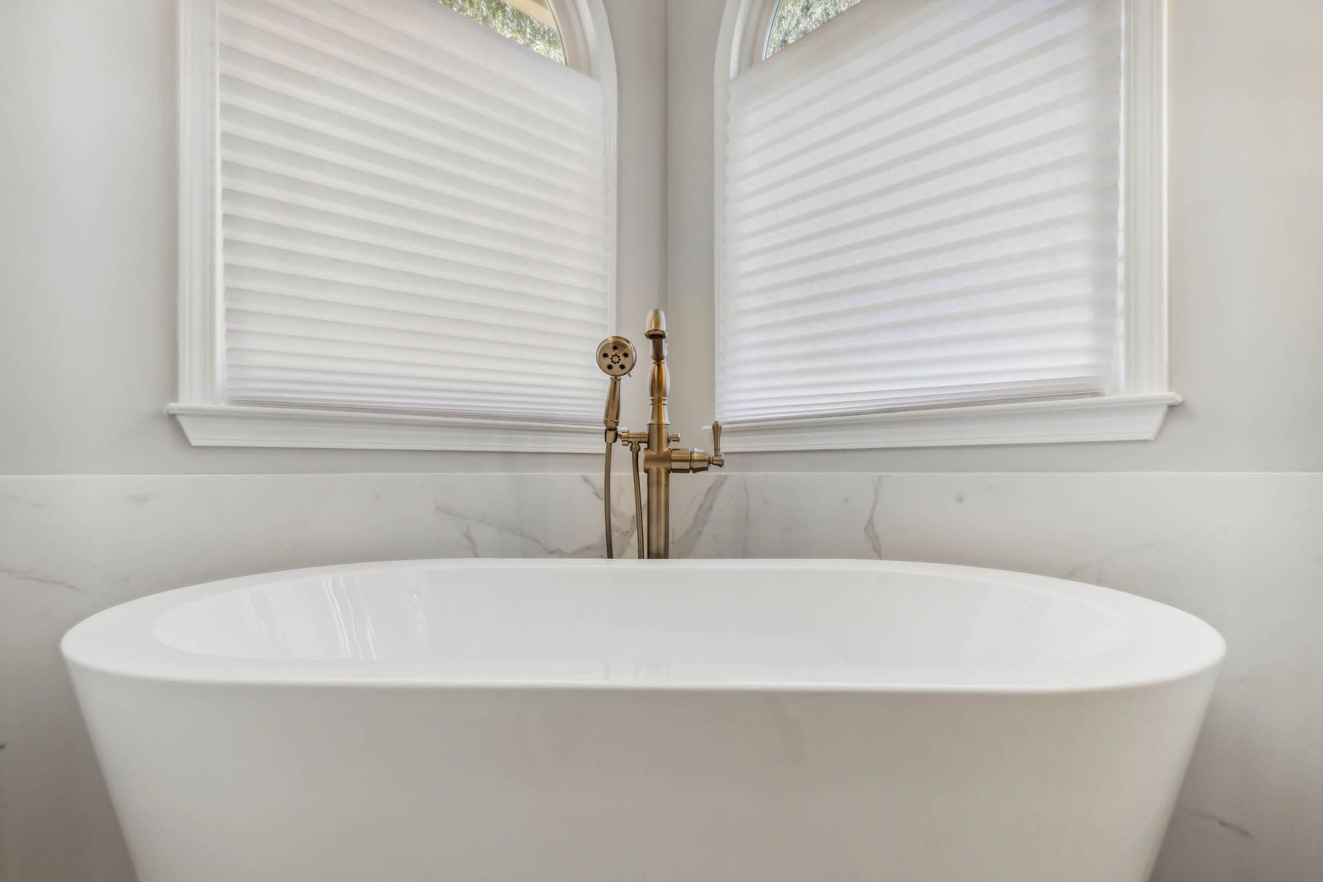A white oval bathtub with gold fixtures sits in front of two windows covered with white pleated blinds.