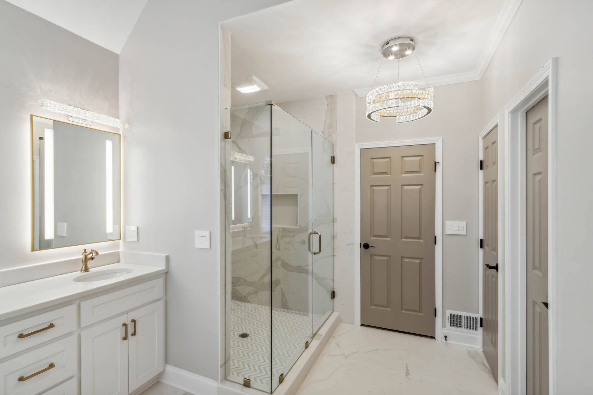 Bright, modern bathroom with a glass shower, vanity, and a gray door leading to the bedroom. Soft gray and white color palette.