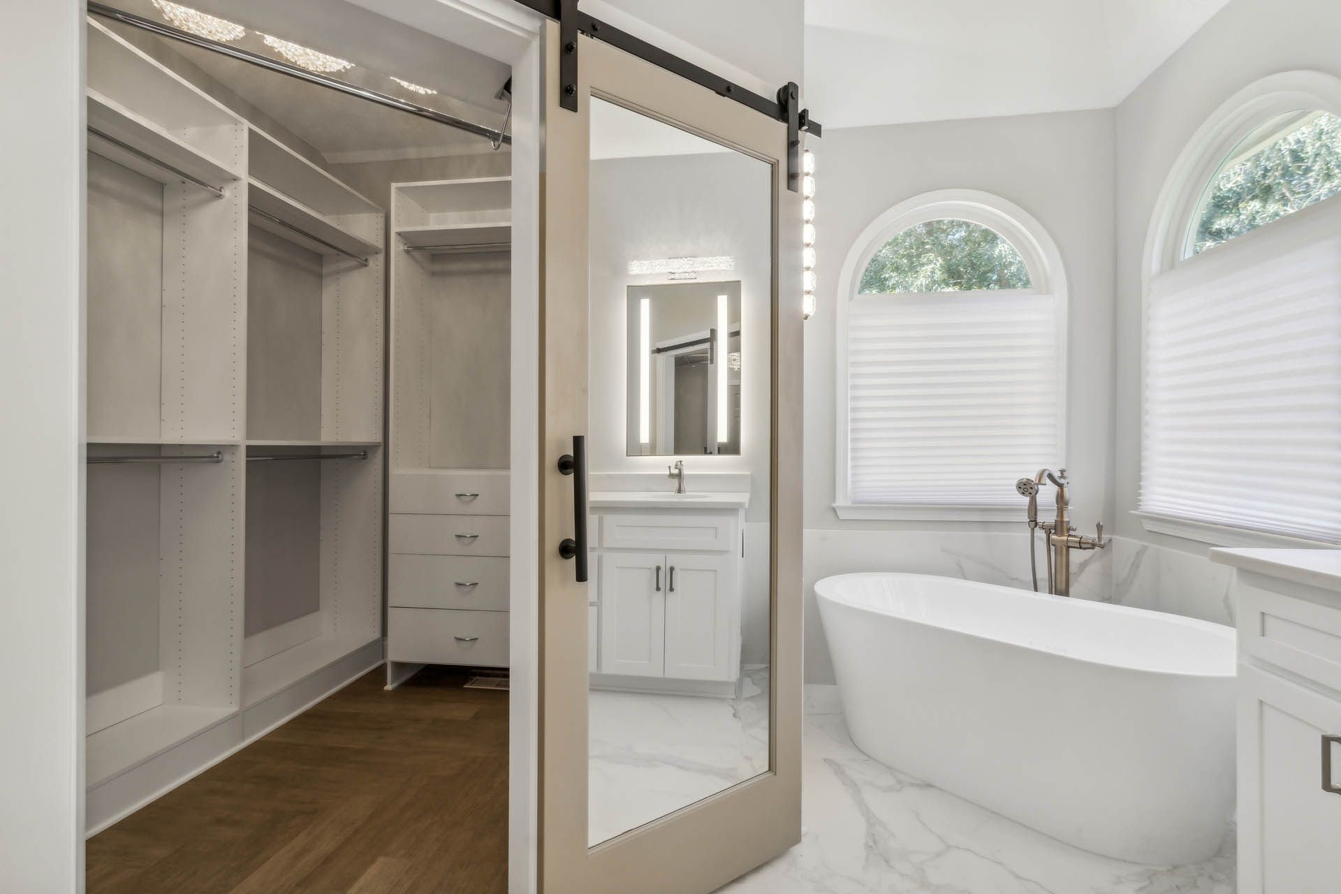 A bright white bathroom with a soaking tub and a connected walk-in closet visible through a sliding glass door.