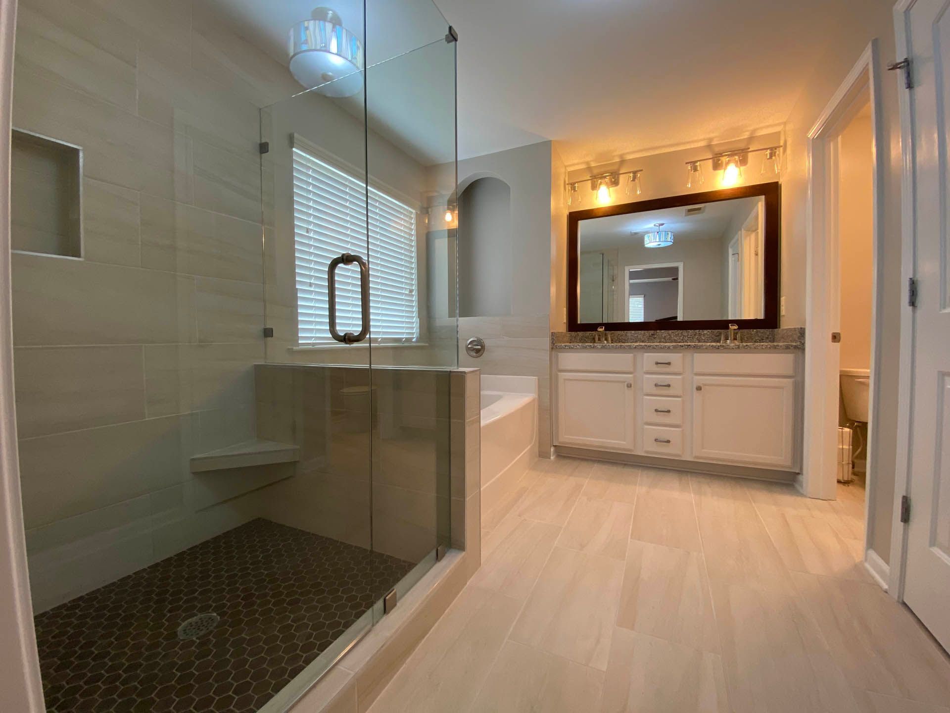 A modern bathroom with a glass shower, white vanity, and light-colored tile flooring. Sunlight streams through a window.