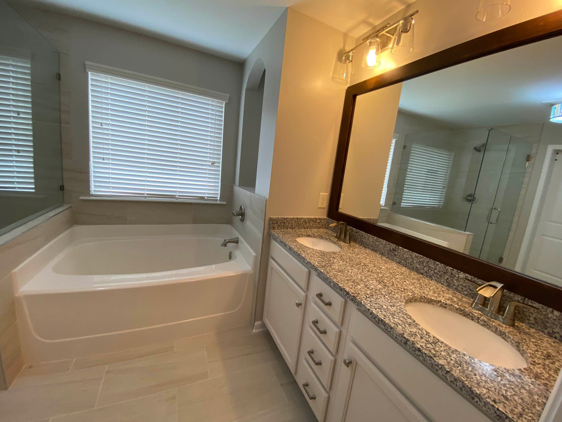 Bathroom with a tub, vanity with granite countertop, large mirror, and window with blinds. The room is light-toned with neutral colors.